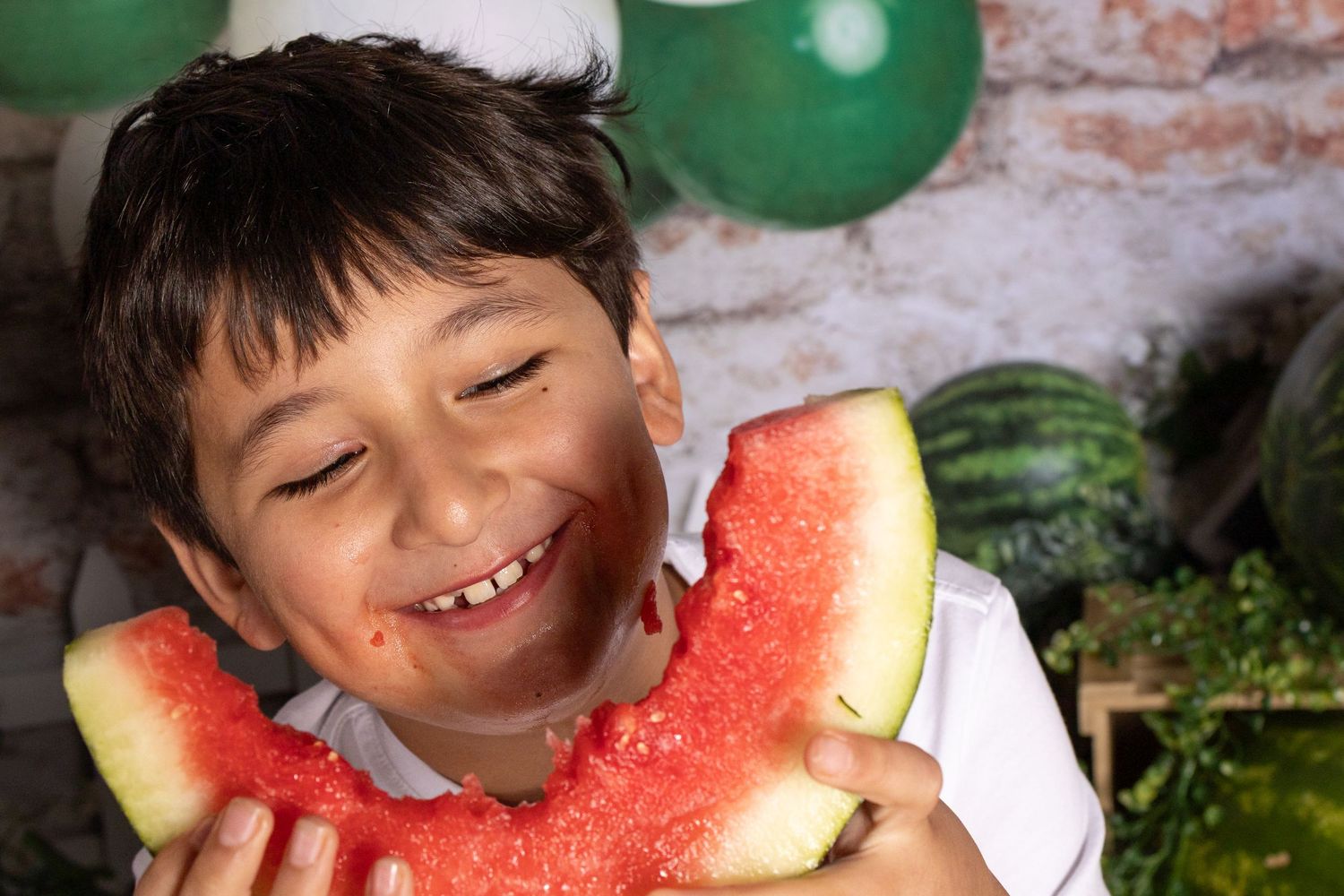 A happy boy smiling while holding a large slice of fresh watermelon, with whole watermelons in the background.