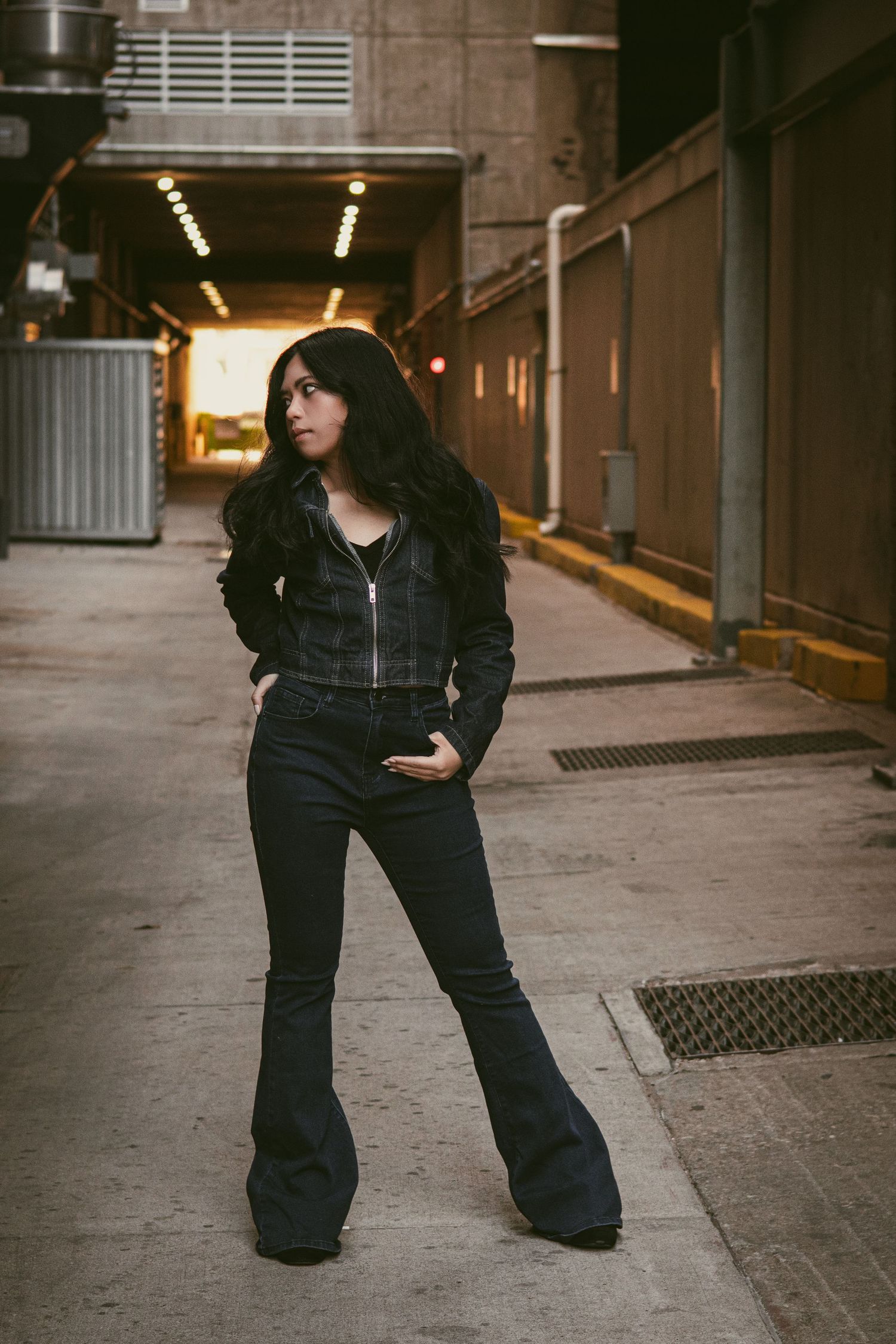 Woman in black flare jeans and jacket posing in urban alley with tunnel lights in background.