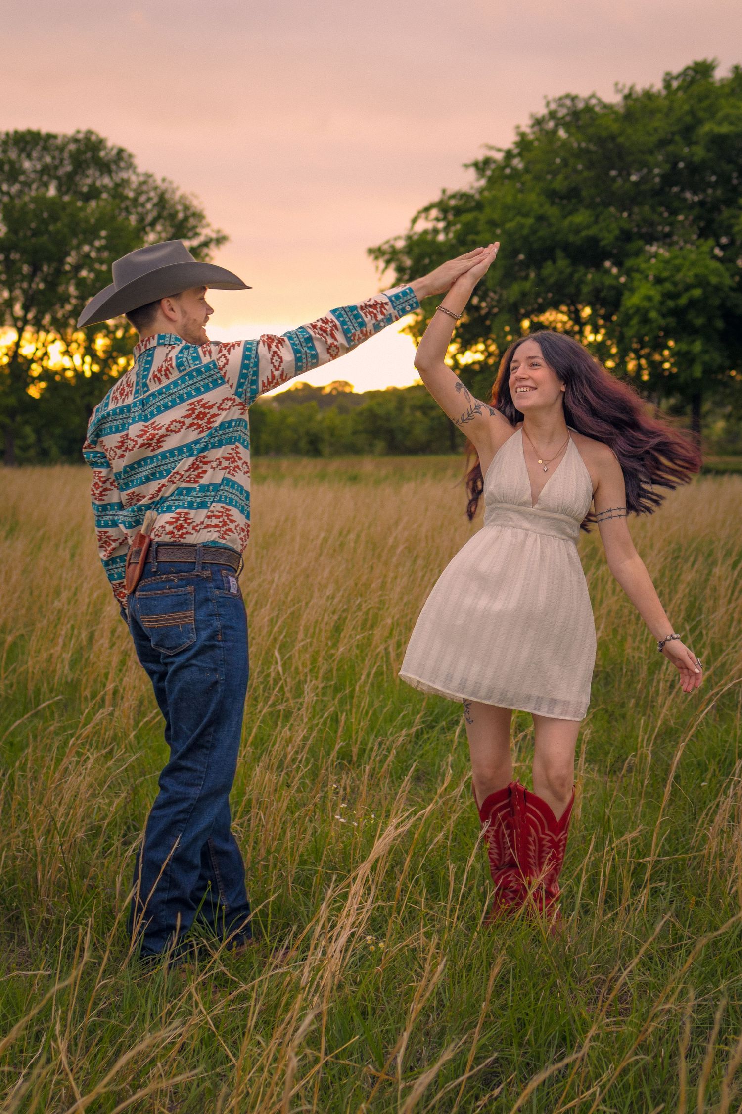 Couple dancing at sunset in a field, man in cowboy hat and western shirt, woman in white dress and red cowboy boots.