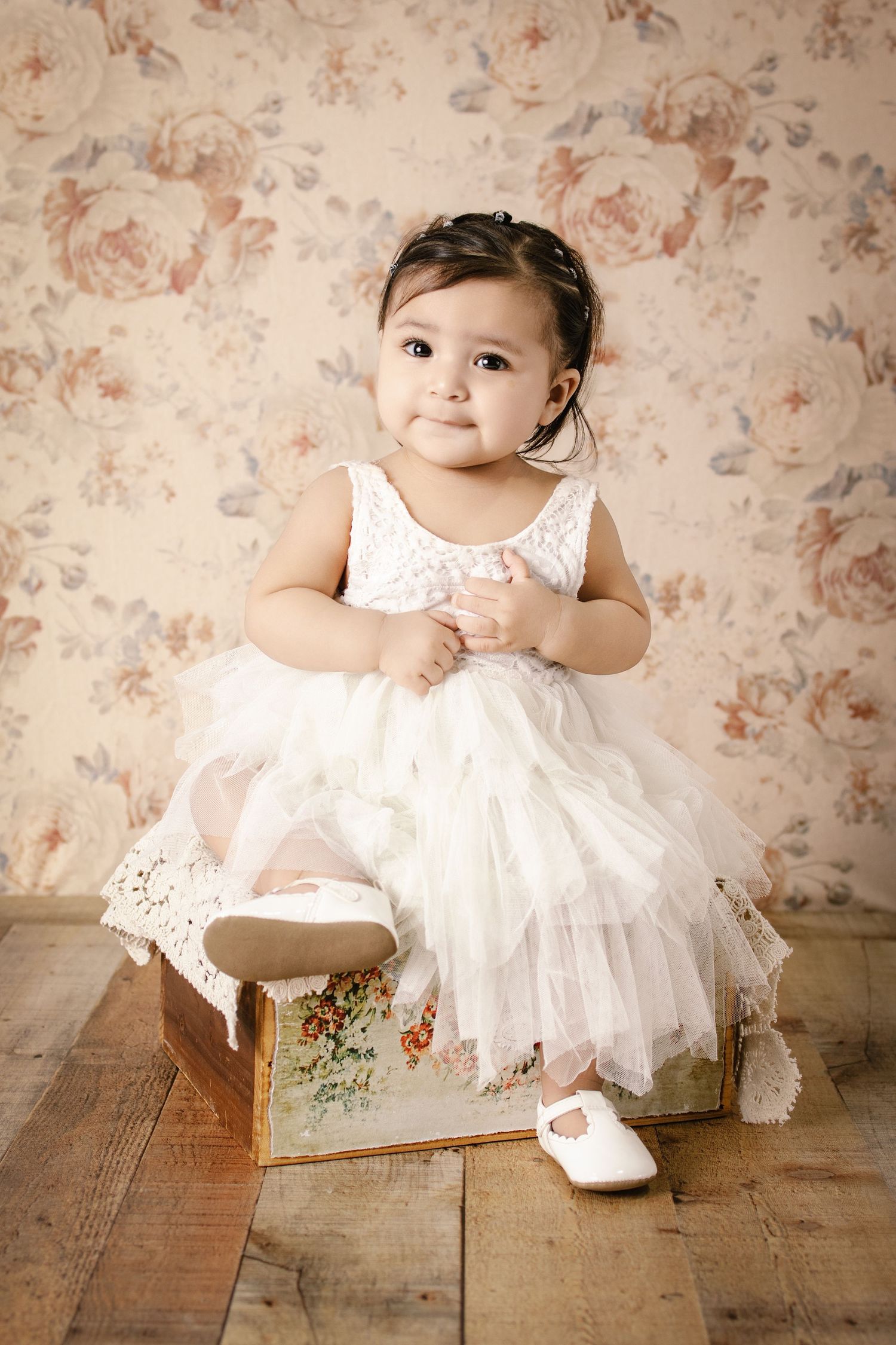 Adorable baby girl in white lace tutu dress sitting on vintage floral box with matching wallpaper background.