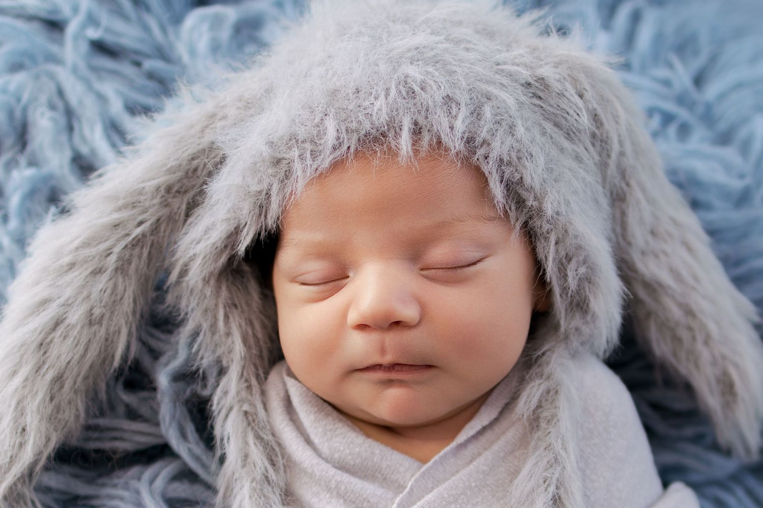 Sleeping newborn baby wearing a soft gray fuzzy bunny ear hat, wrapped in a light gray swaddle on a blue fur blanket.