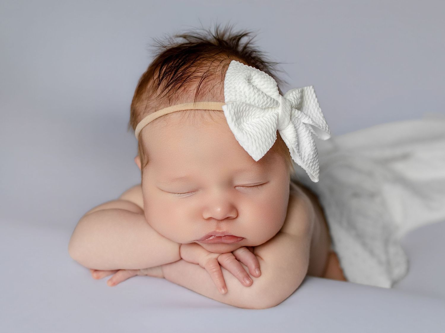 Peaceful newborn baby sleeping with chin resting on hands, wearing a white bow headband on a soft gray background.