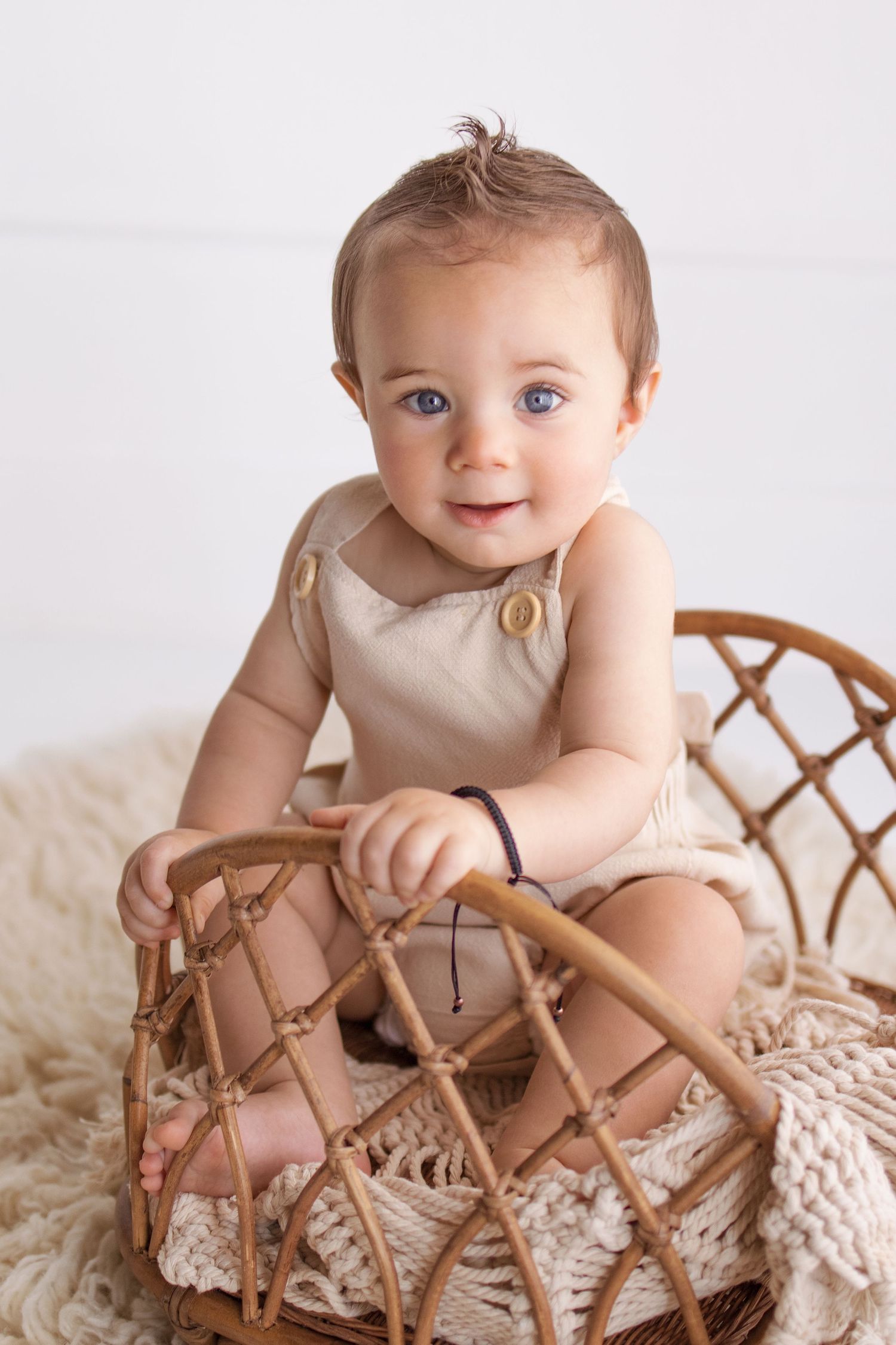 Smiling baby with blue eyes sitting in a wicker basket wearing a beige romper on a cream fluffy rug.