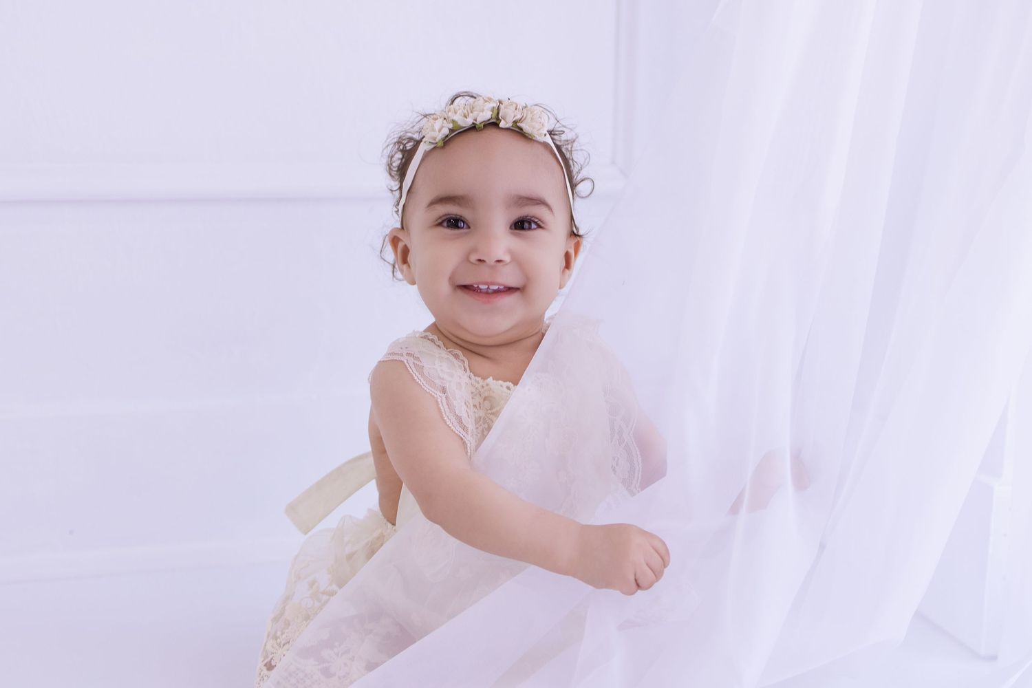 Smiling baby girl in white lace dress and floral headband poses by sheer curtains in bright white studio.