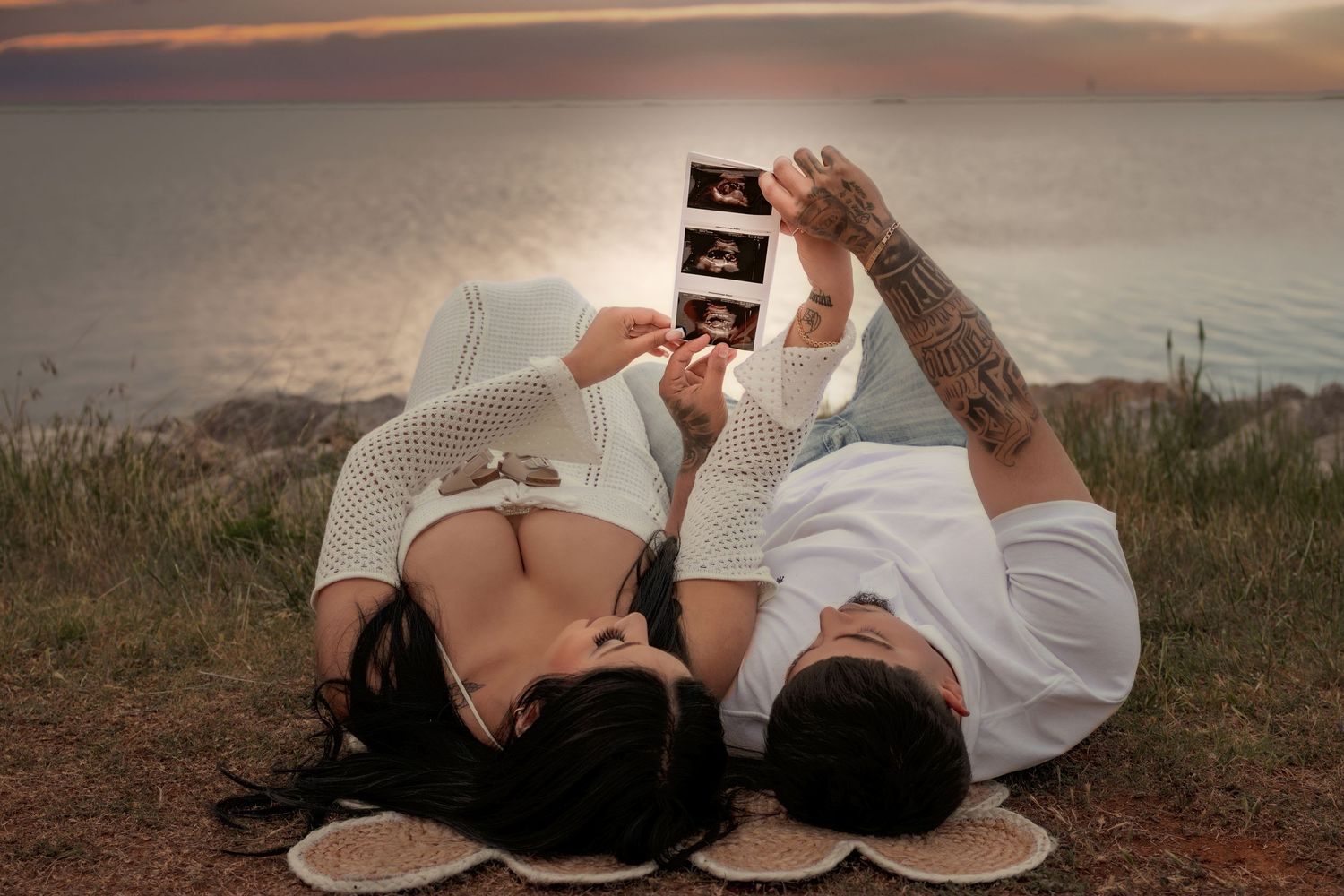 Couple lying on grass holding ultrasound photos against sunset ocean backdrop, announcing pregnancy outdoors.