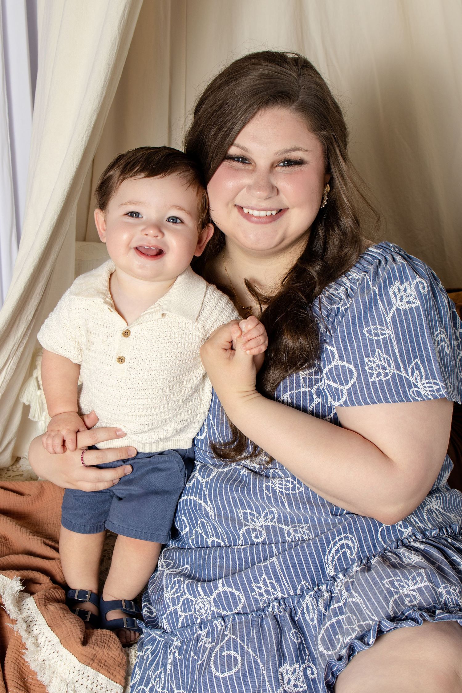A smiling young woman in a blue floral dress poses with a happy baby boy in a cream top and blue shorts.
