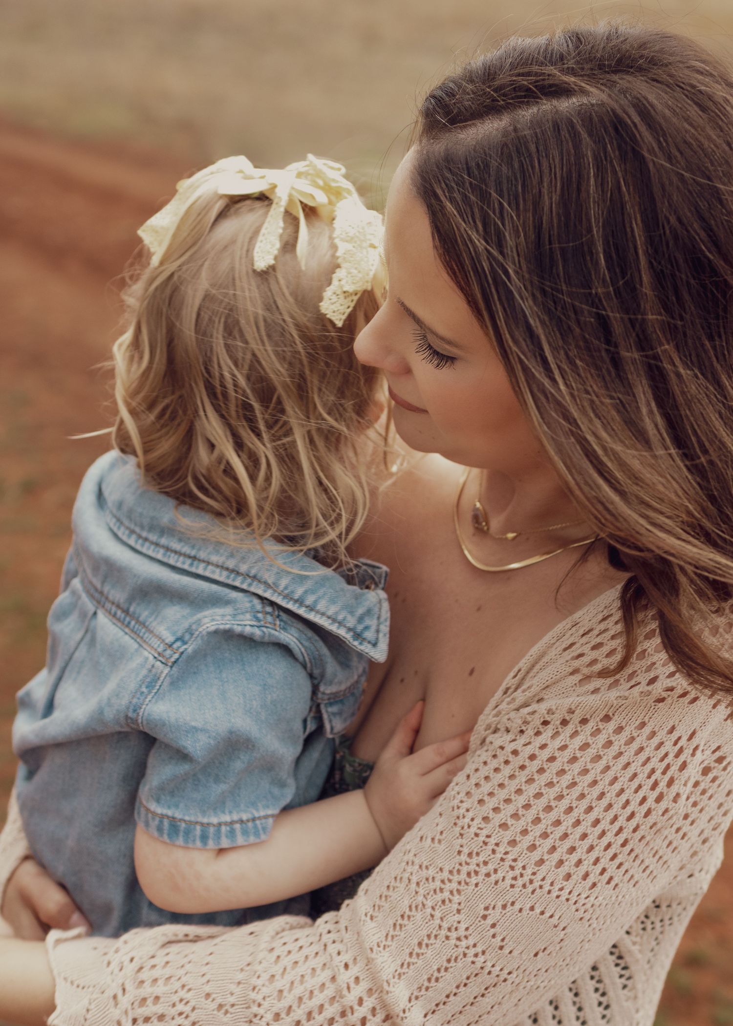 A mother in a cream knit top tenderly holds her toddler girl in a denim jacket with a floral bow outdoors.