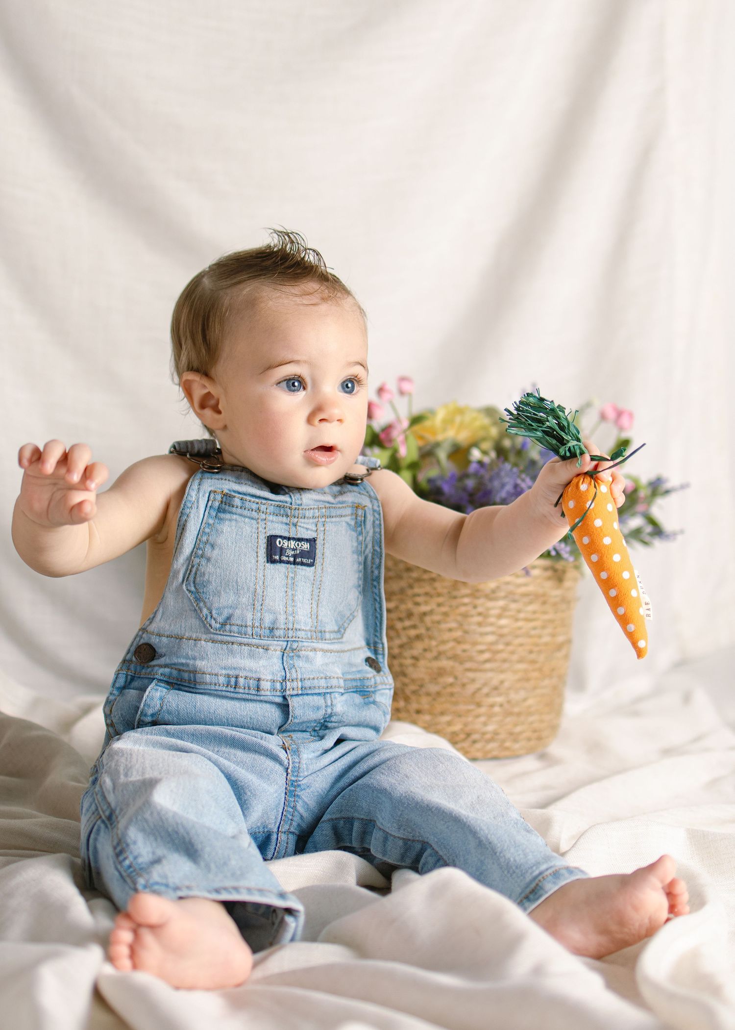 Curious baby in denim overalls holds a polka dot toy carrot during an Easter-themed photo shoot.