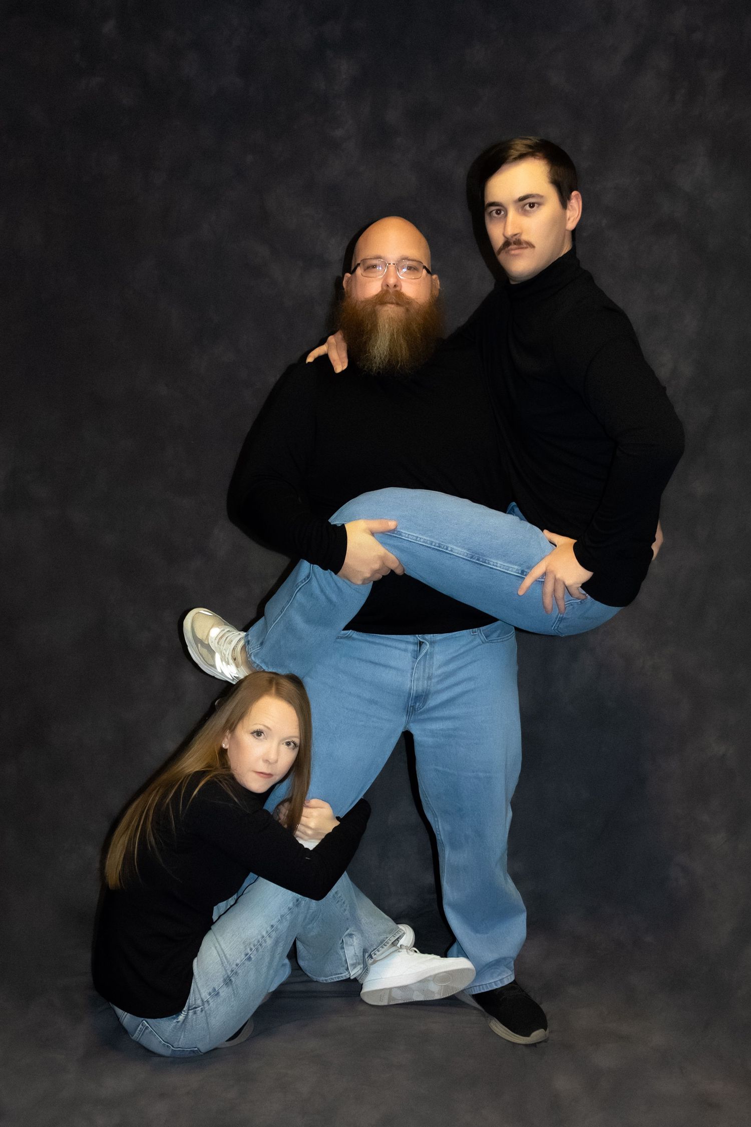 Three people in black turtlenecks and jeans pose humorously in a studio, with a bearded man holding another mans legs up.