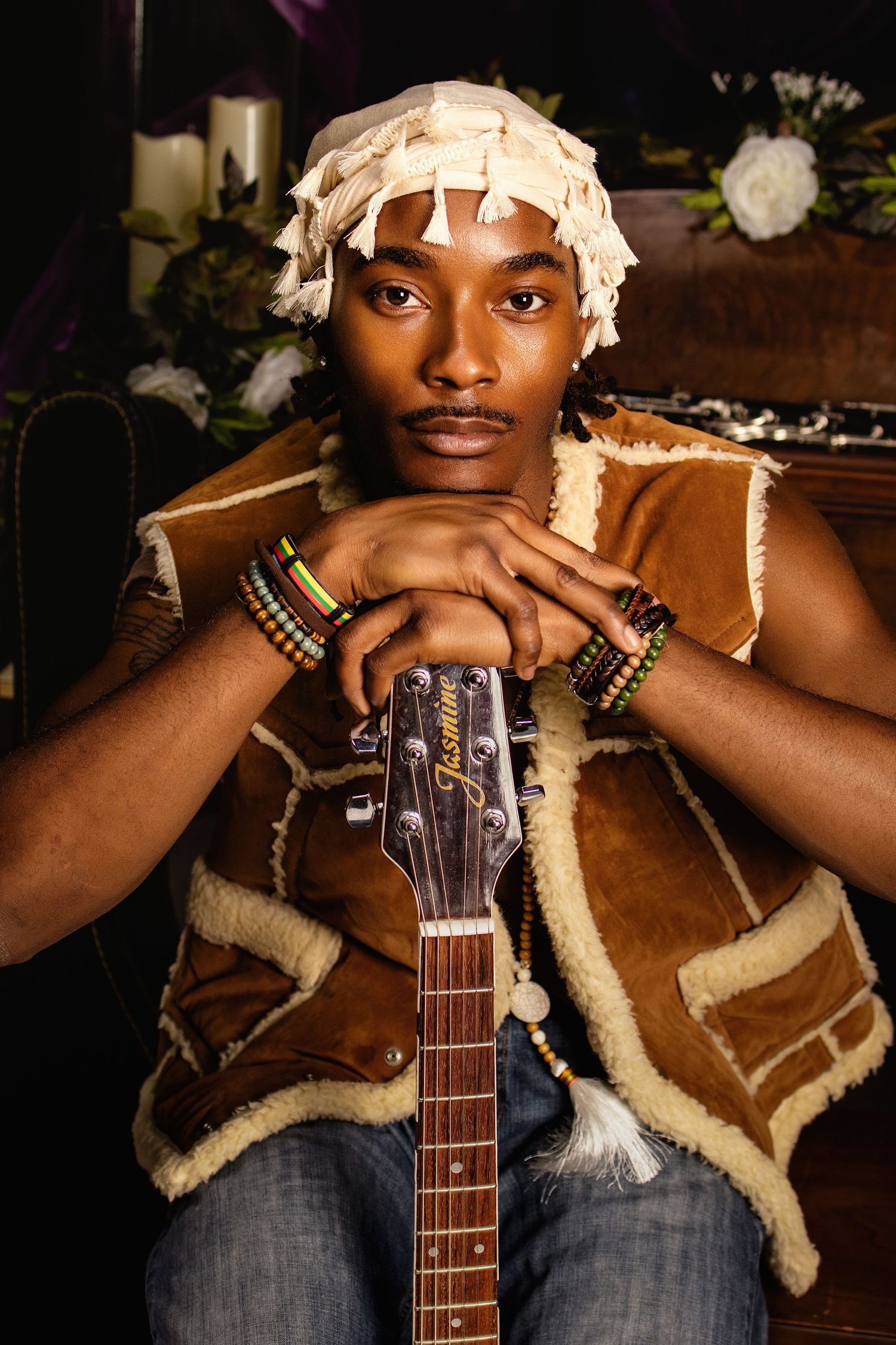 Young Black musician resting chin on Jasmine acoustic guitar neck, wearing shearling vest and colorful bracelets with wrapped head covering.