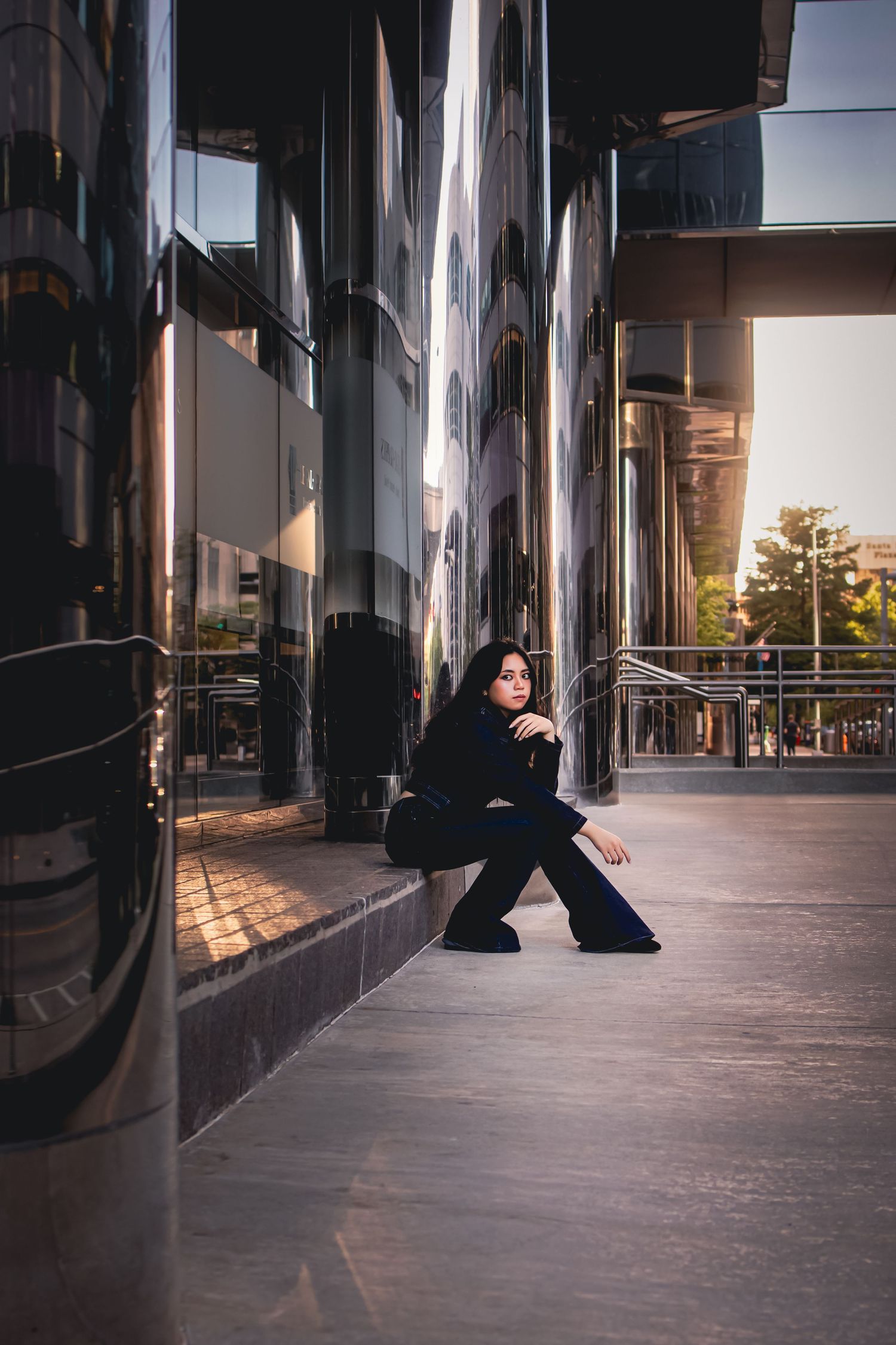 A woman in all-black outfit sits gracefully against a reflective modern building facade at dusk.