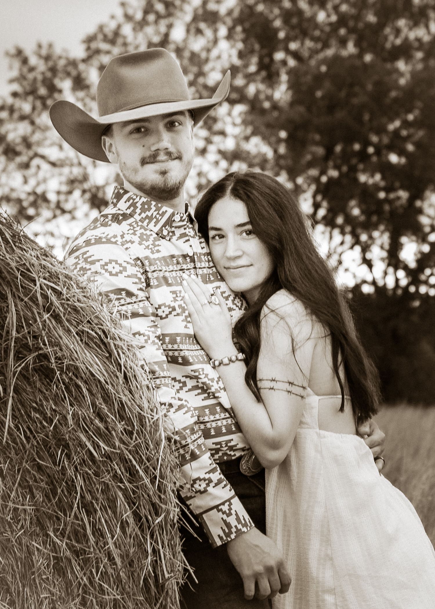 Couple poses romantically by a hay bale in sepia tone, man in cowboy hat and patterned shirt with woman in white dress.