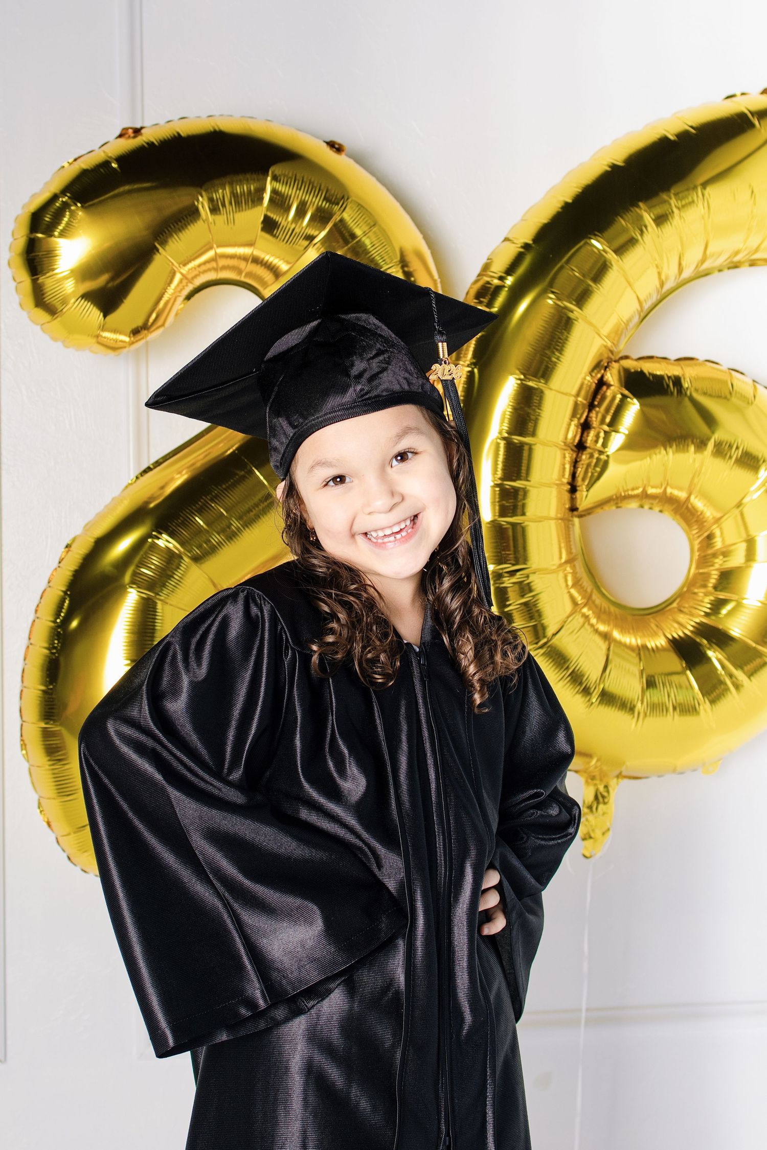 Young girl in black graduation cap and gown smiling proudly in front of gold number 26 balloons.
