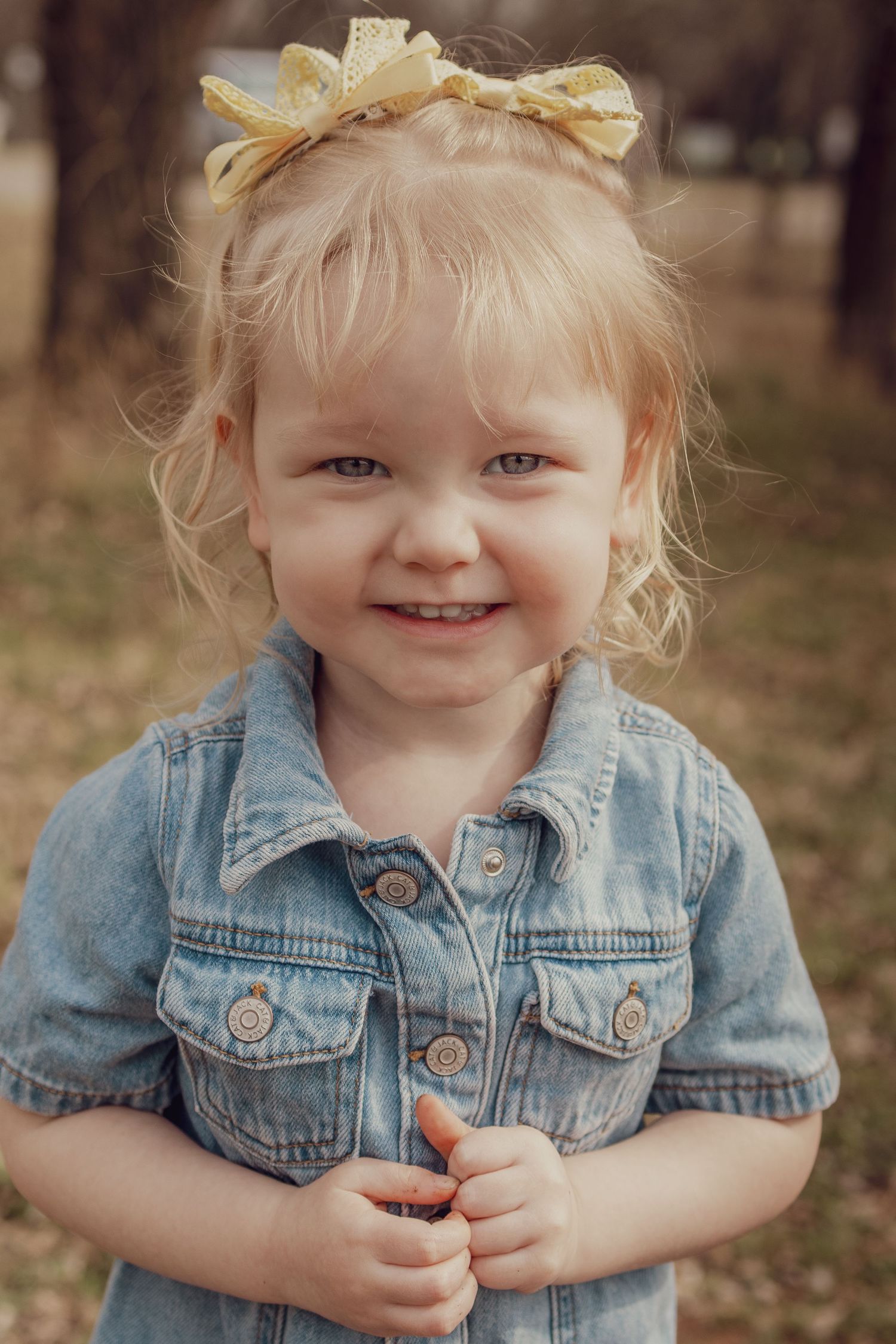 Smiling toddler girl with blonde hair and yellow bow wearing denim dress outdoors.
