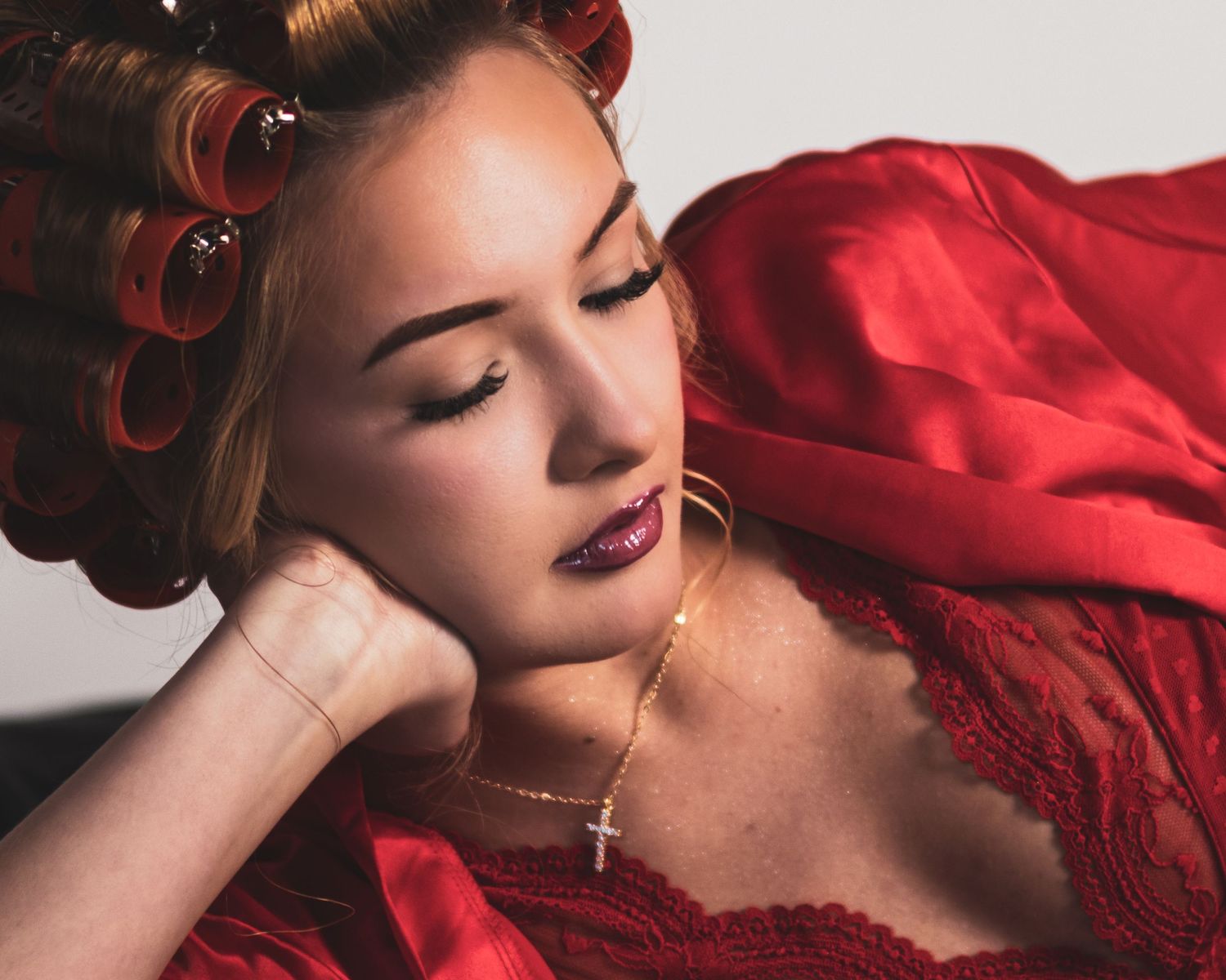 Woman with red hair rollers and dark lips wearing red lace, gold cross necklace, eyes closed in elegant pose.