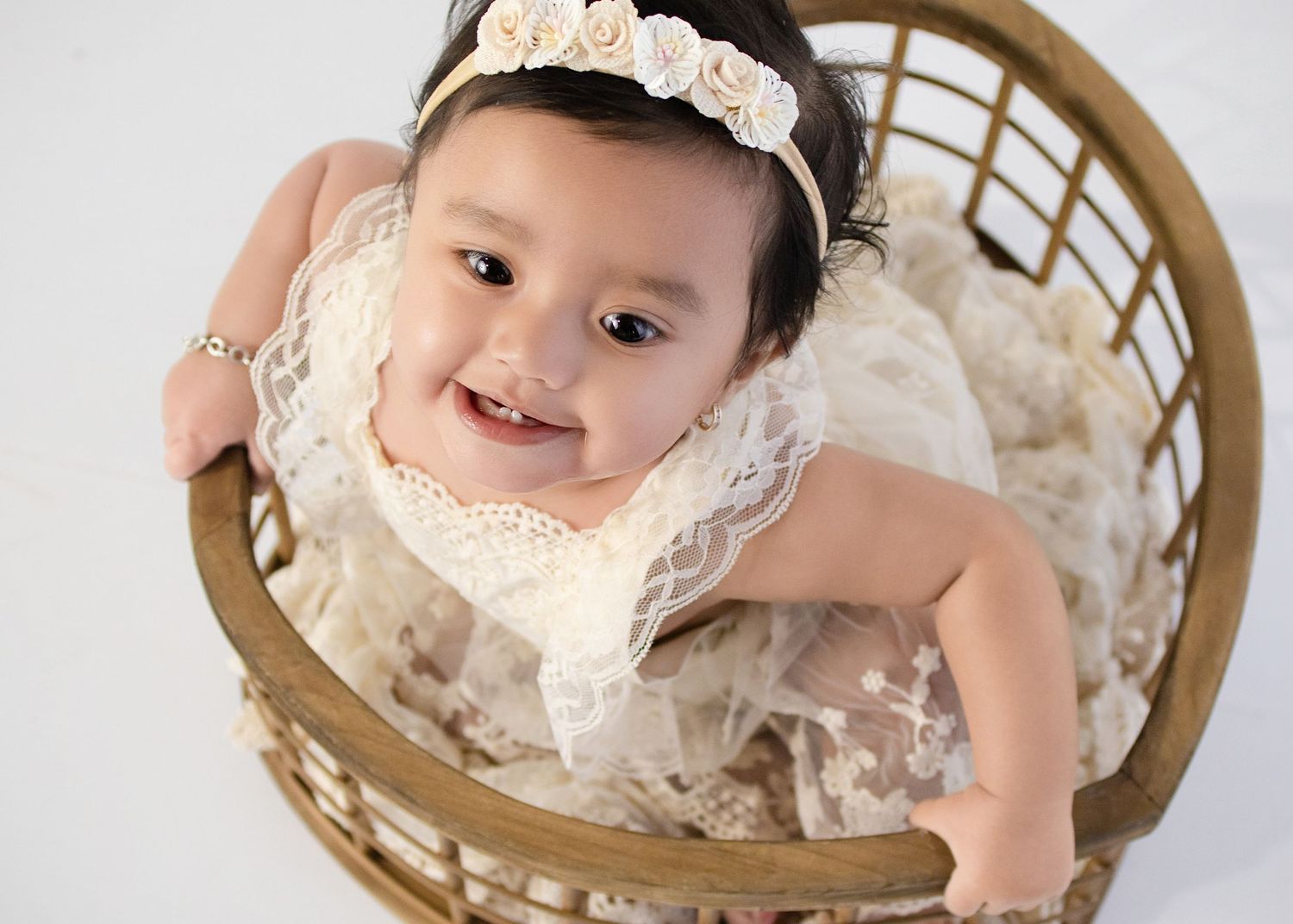 Smiling baby girl in lace dress and floral headband sitting in a wicker basket on white background.