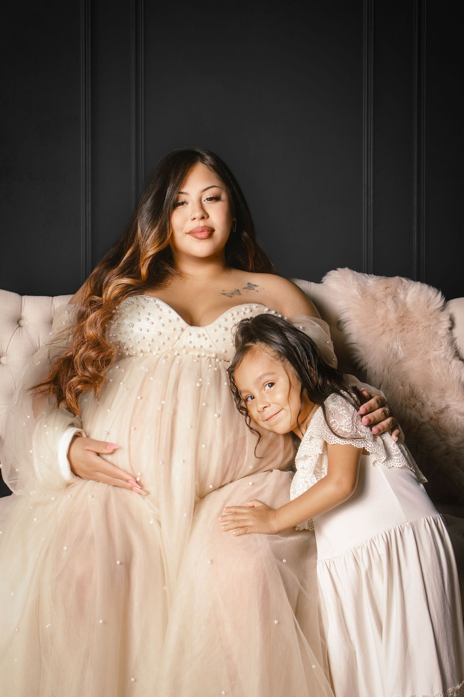 Pregnant woman in pearl gown poses with young daughter in white dress on elegant sofa against dark wall.
