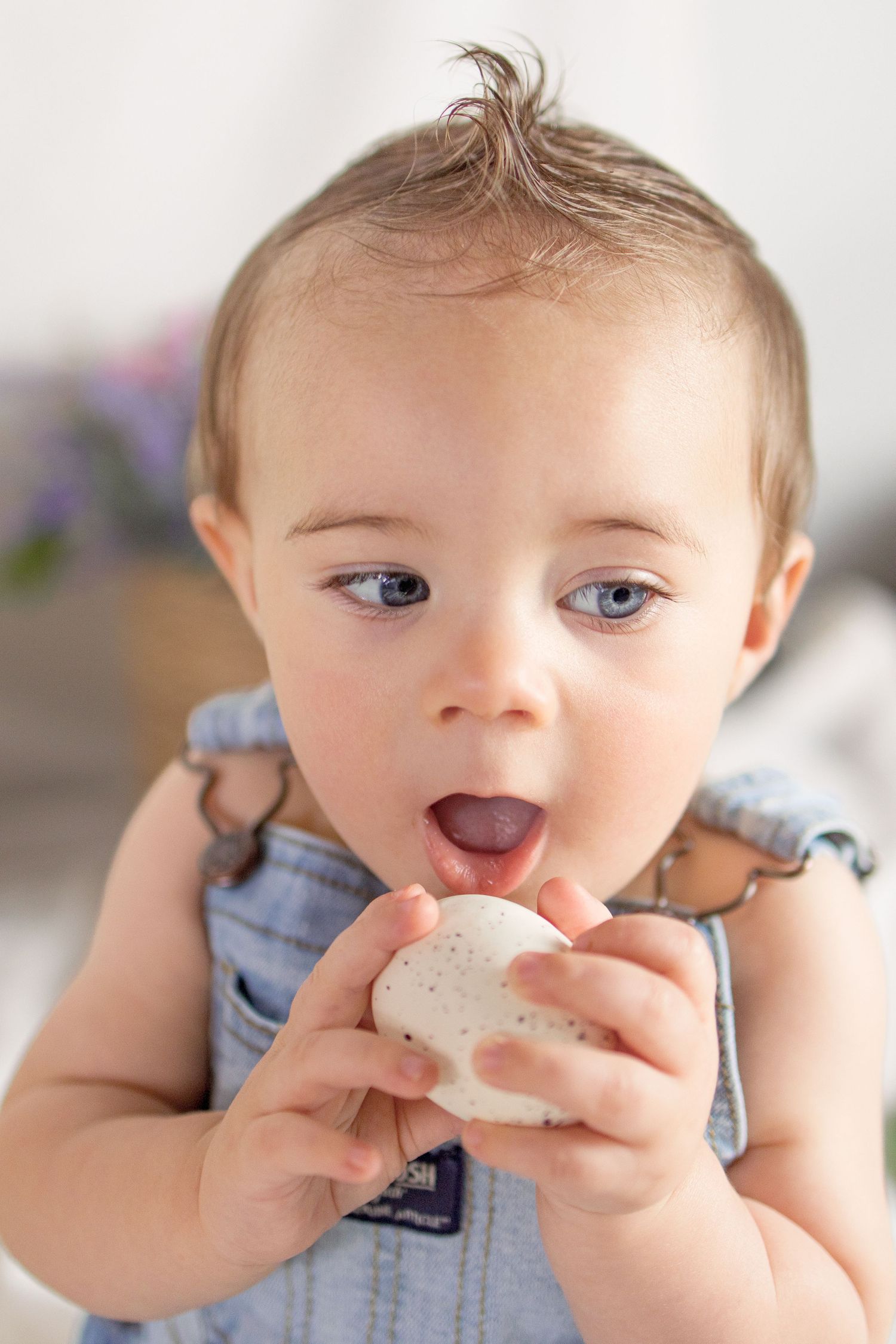 Cute baby with blue eyes in denim overalls licking a white Easter egg.