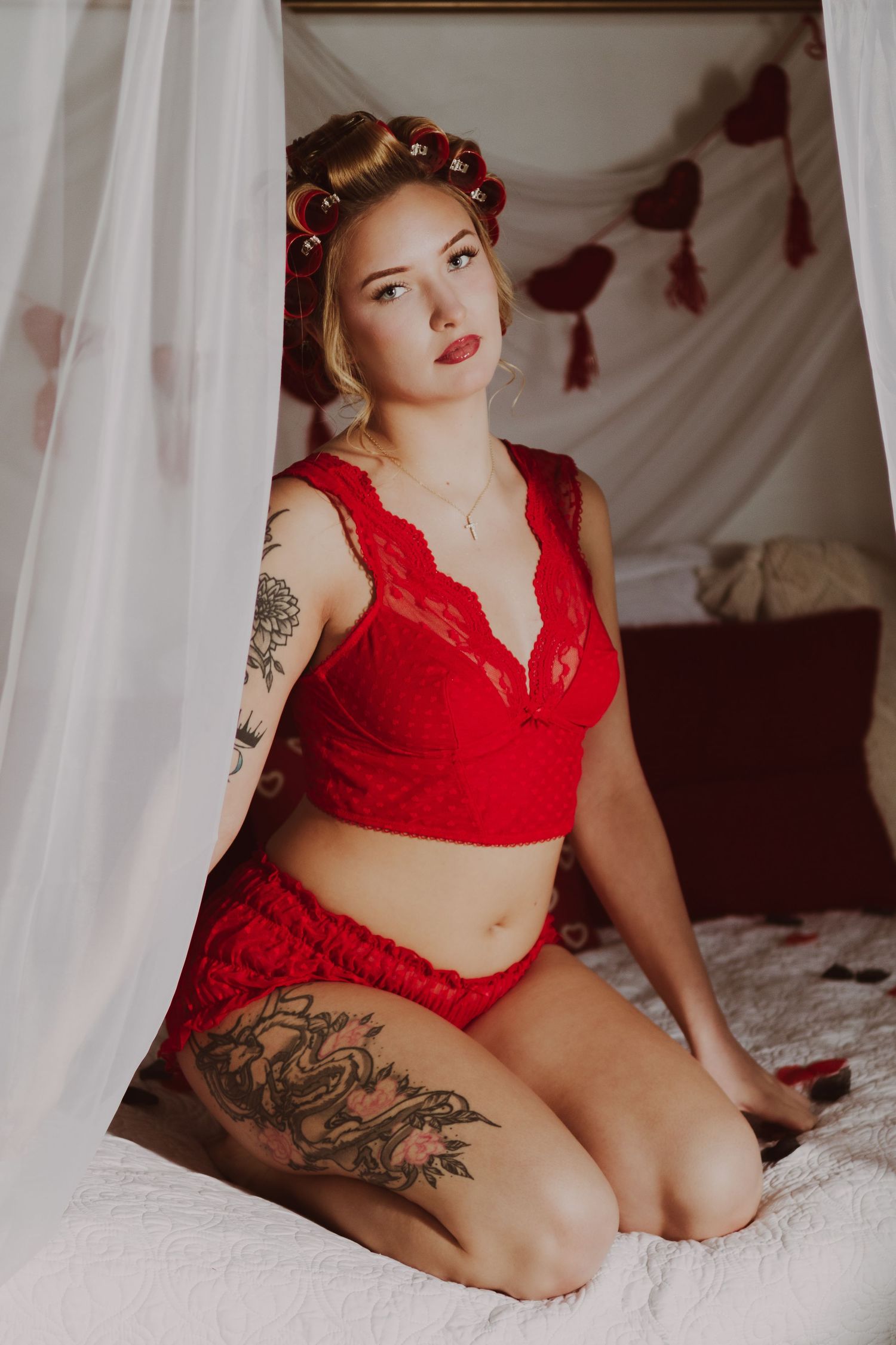 Woman in red lace lingerie with tattoos and hair rollers kneeling on a bed with romantic decor.