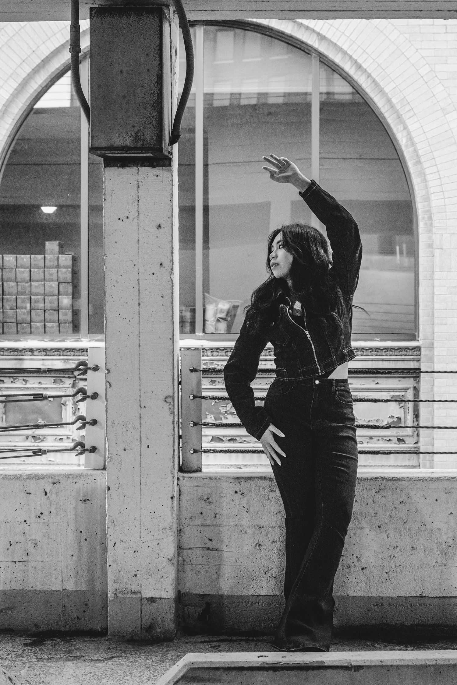Black and white photo of woman in dark outfit posing with raised arm near arched window in urban setting.