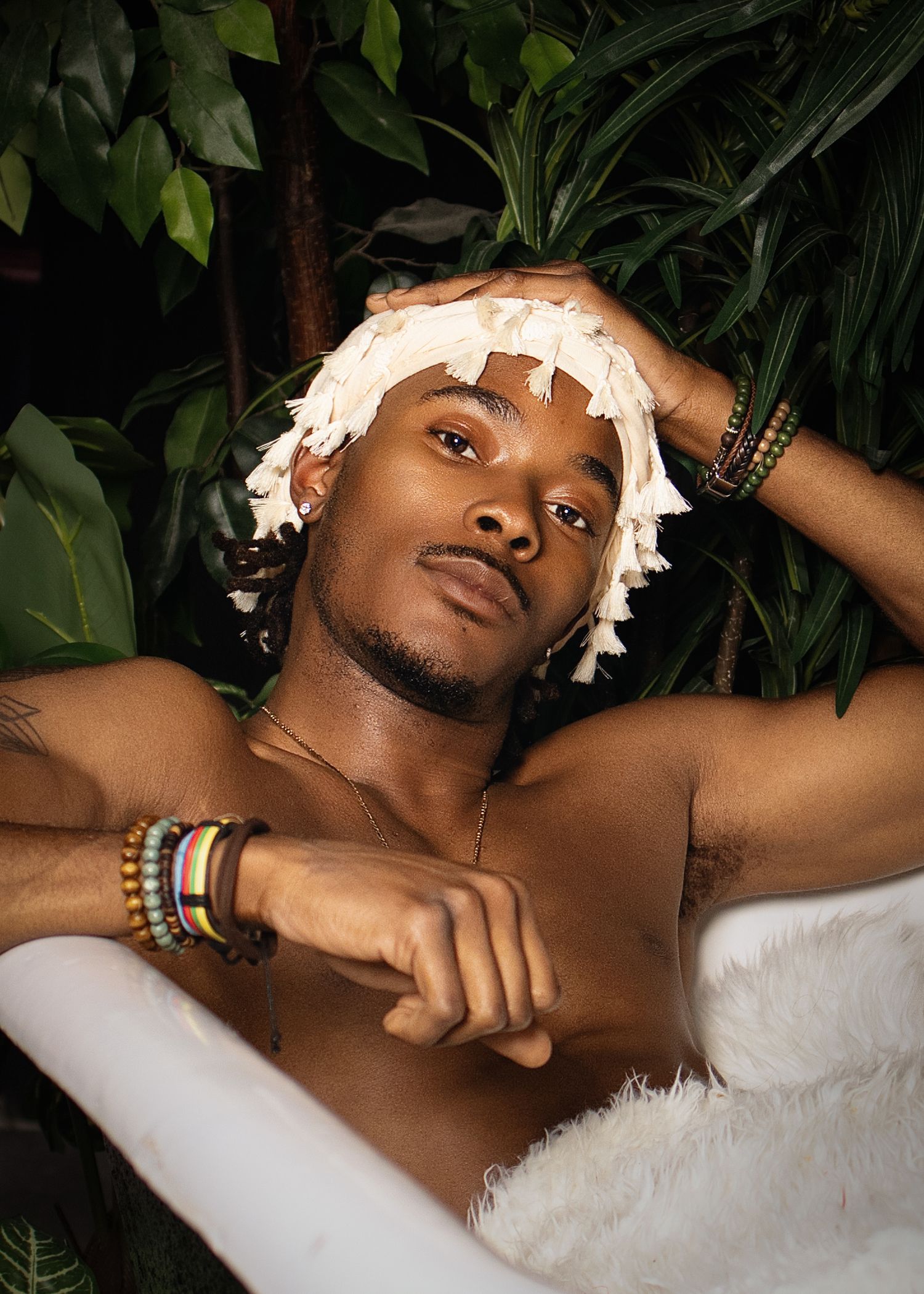 Shirtless Black man with colorful bracelets and white head wrap relaxes in bathtub with tropical plants.