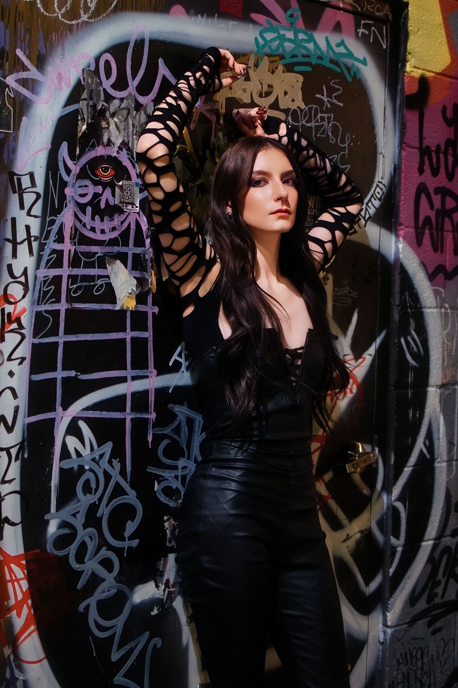 Woman in black outfit with shredded sleeves posing against colorful graffiti wall.