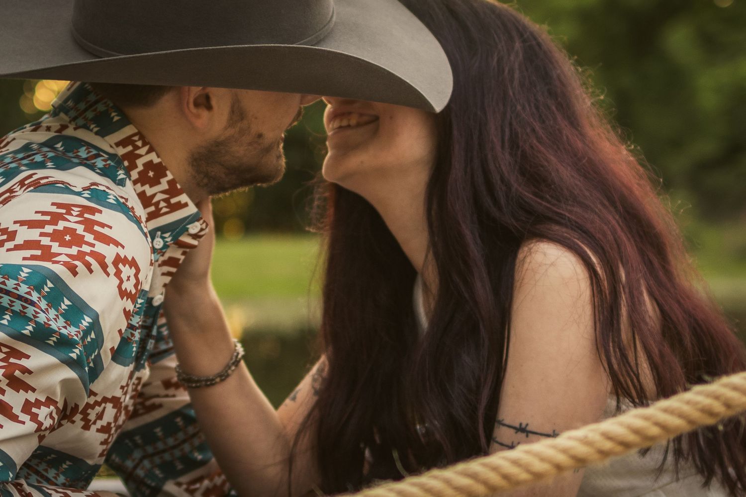 Couple in cowboy hats sharing a romantic moment outdoors, wearing western attire near a rope fence.