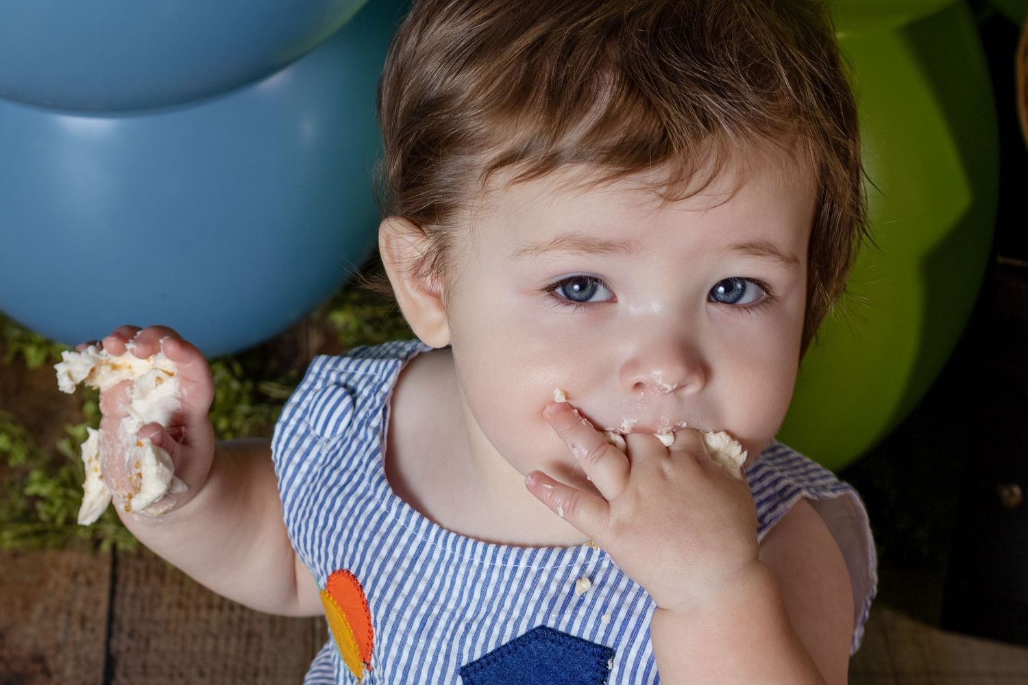 Toddler with blue eyes eating birthday cake with hands at a party with colorful balloons in background.