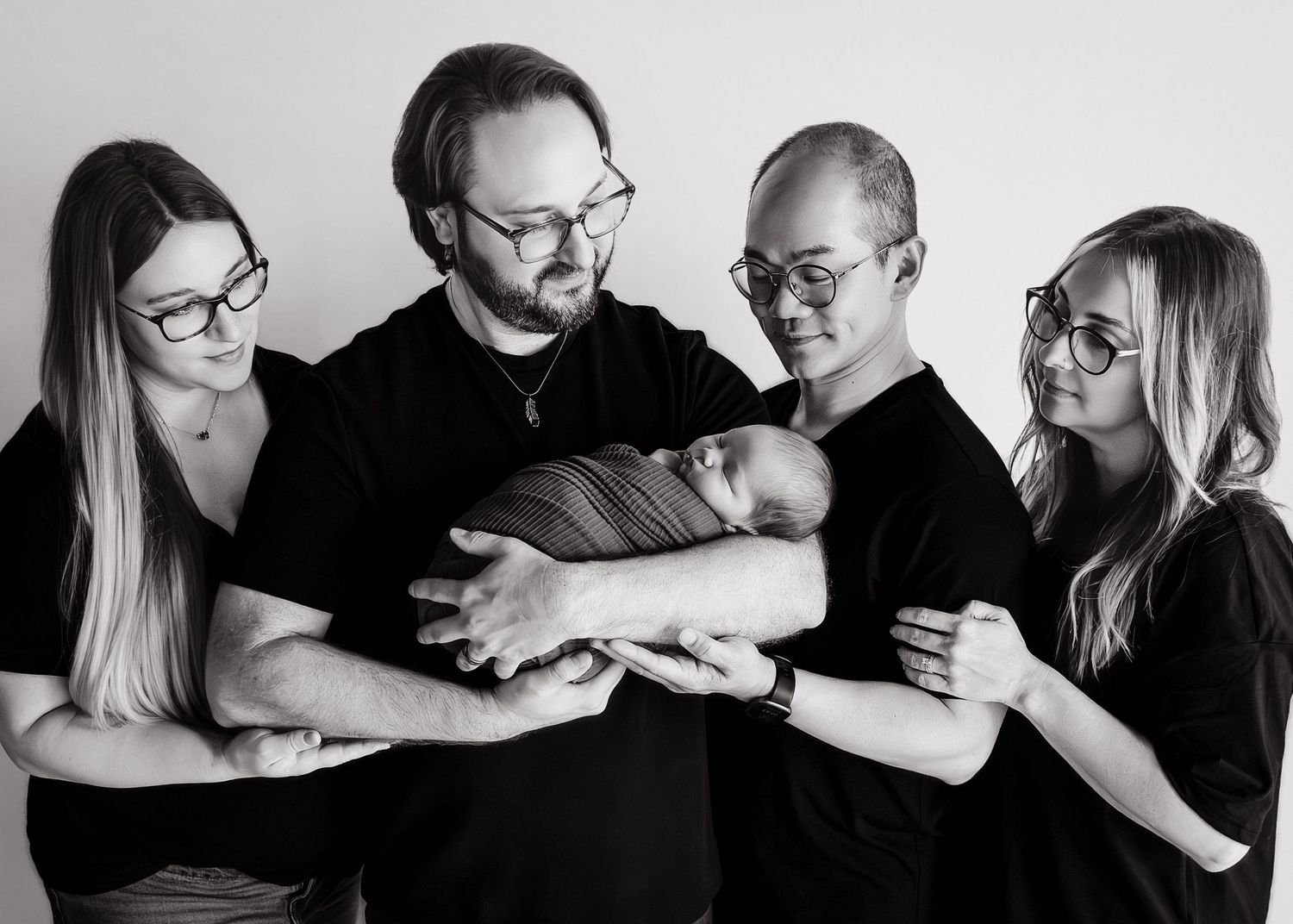 Black and white family portrait of five adults in matching black tops holding a swaddled newborn baby.