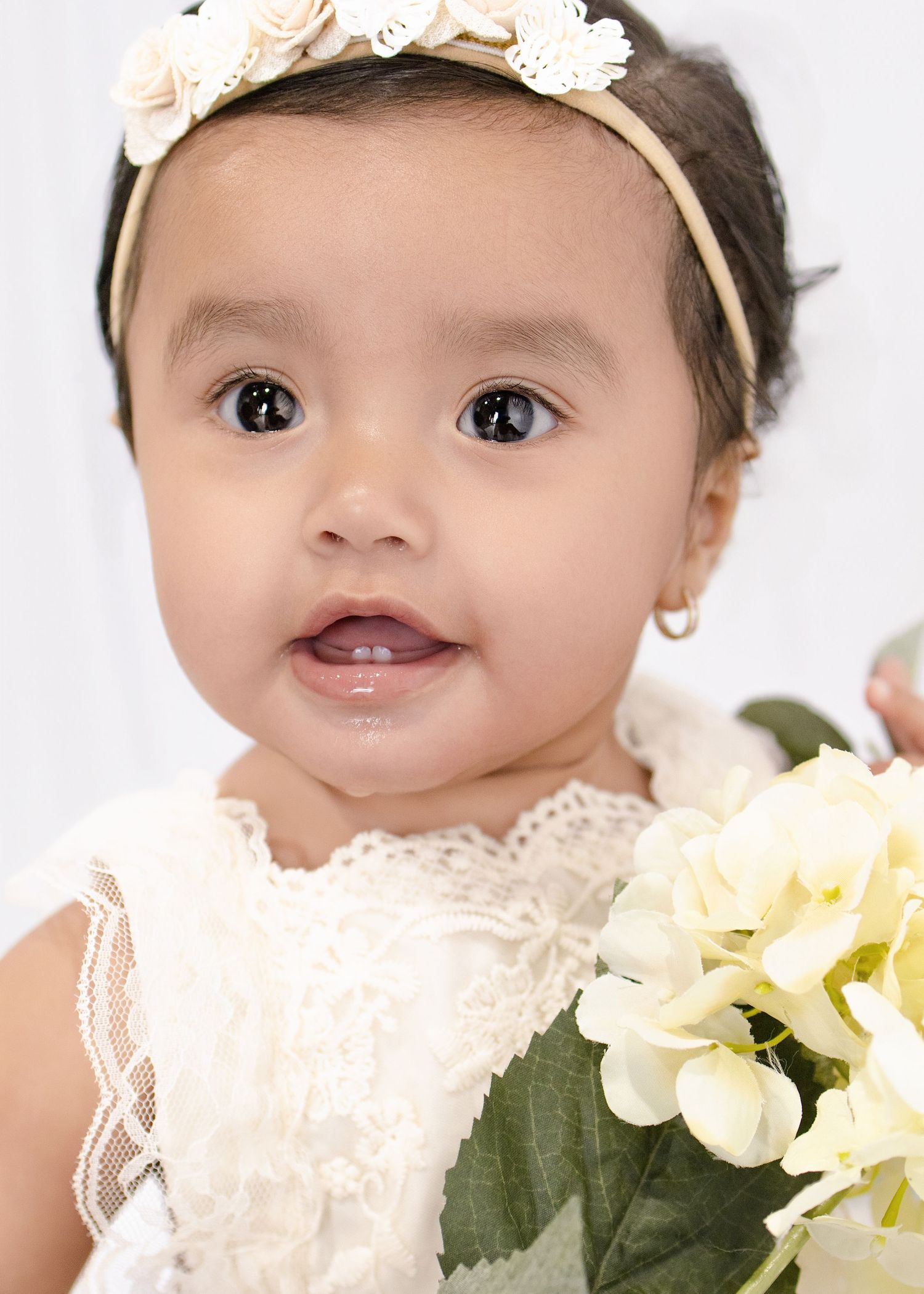 Baby girl in white lace dress and floral headband holds cream hydrangeas, wearing gold hoop earrings.