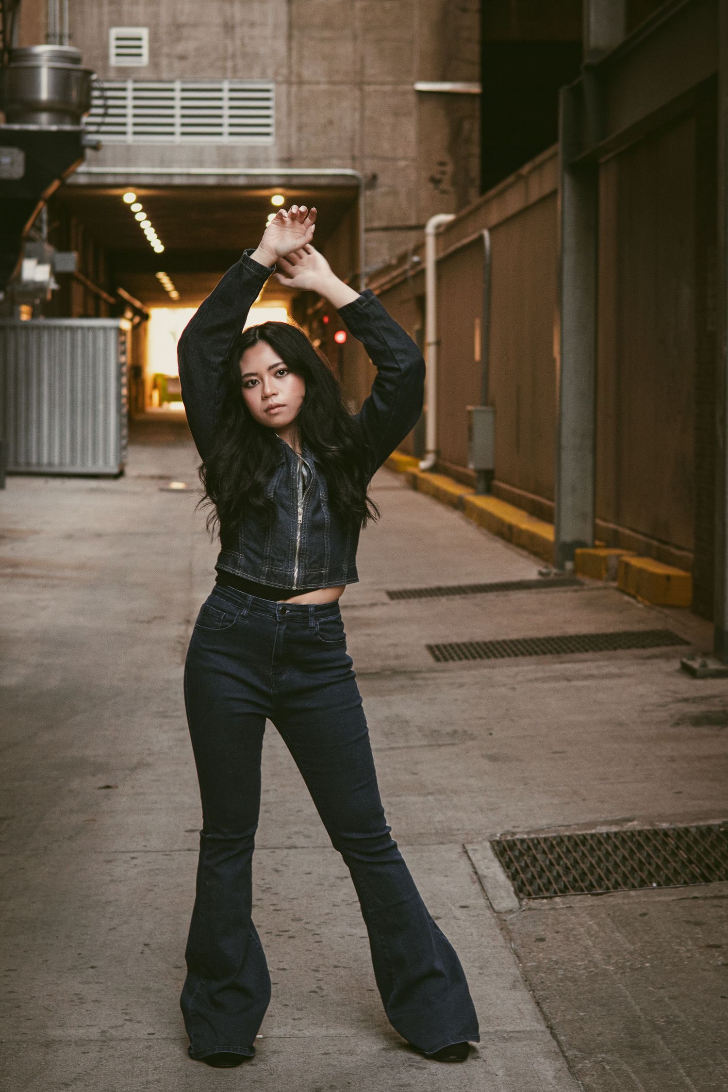Young woman in cropped jacket and flare jeans poses with arms raised in urban alleyway.