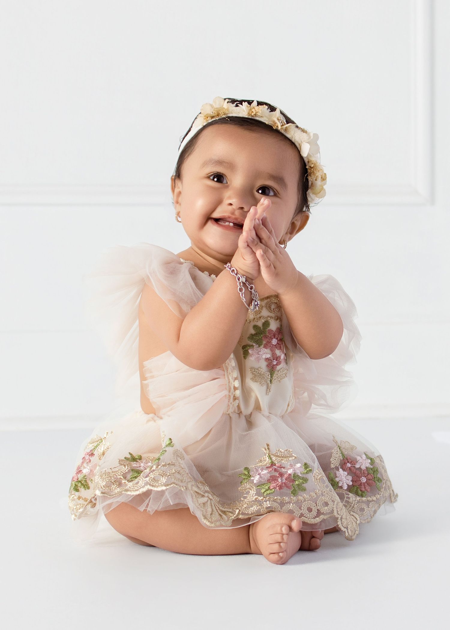 Smiling baby girl in floral embroidered tulle dress and flower headband, clapping hands, seated on white background.