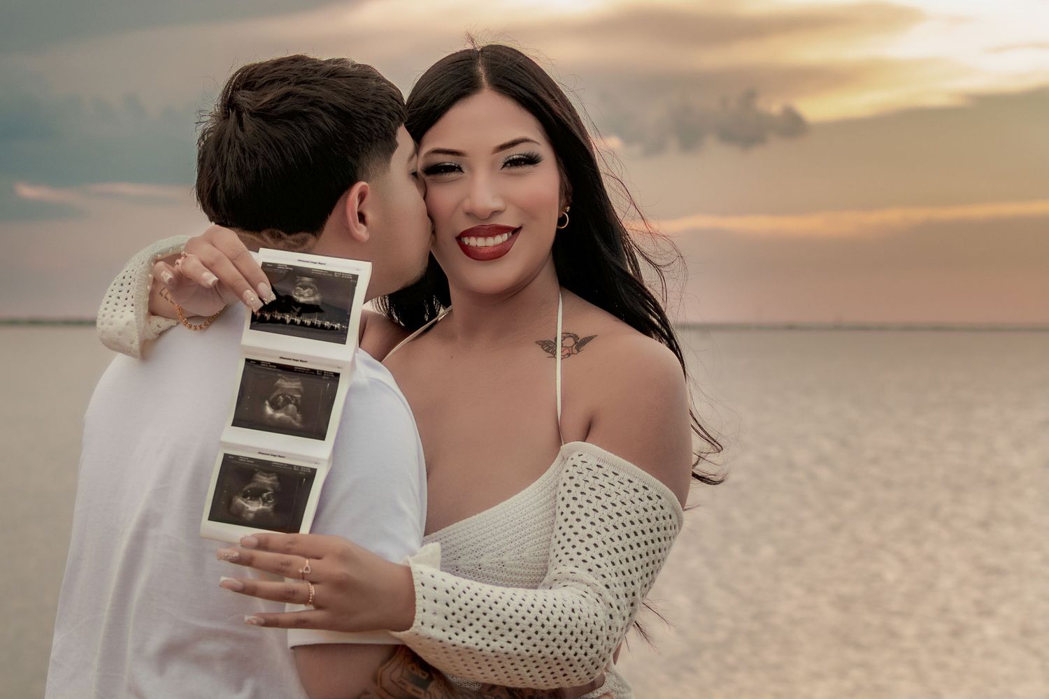 Couple sharing a tender moment on a beach at sunset, holding ultrasound photos for a pregnancy announcement.