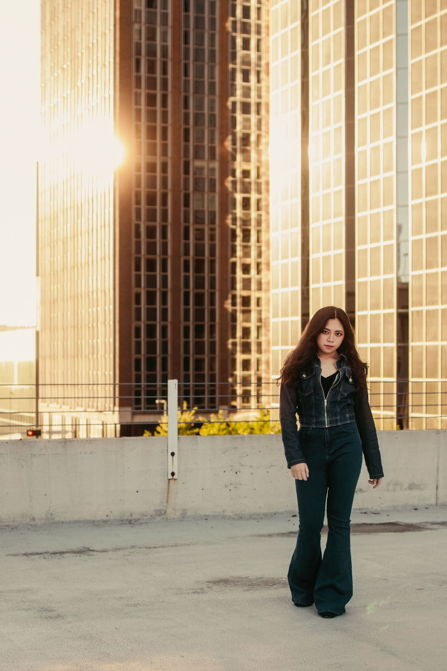 Young woman in black jacket and flare jeans poses on rooftop with golden sunset and city skyscrapers behind her.