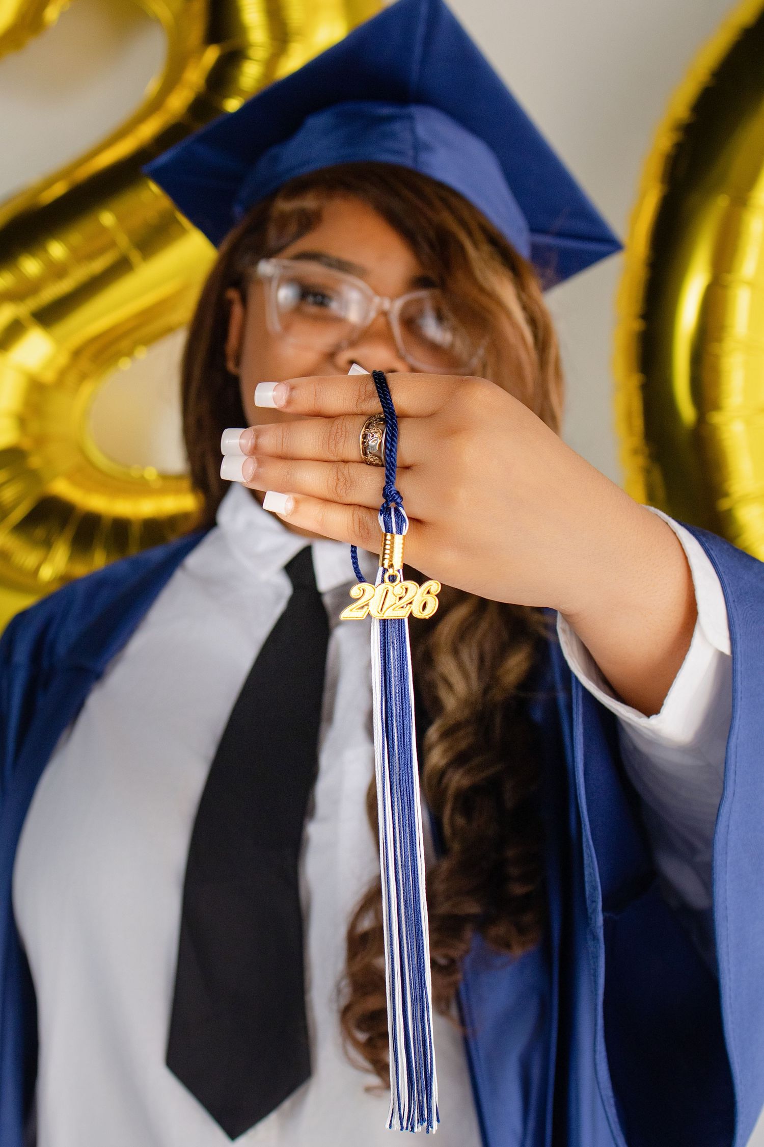 Graduate in blue cap and gown holds 2026 tassel toward camera, with gold number balloons in background.