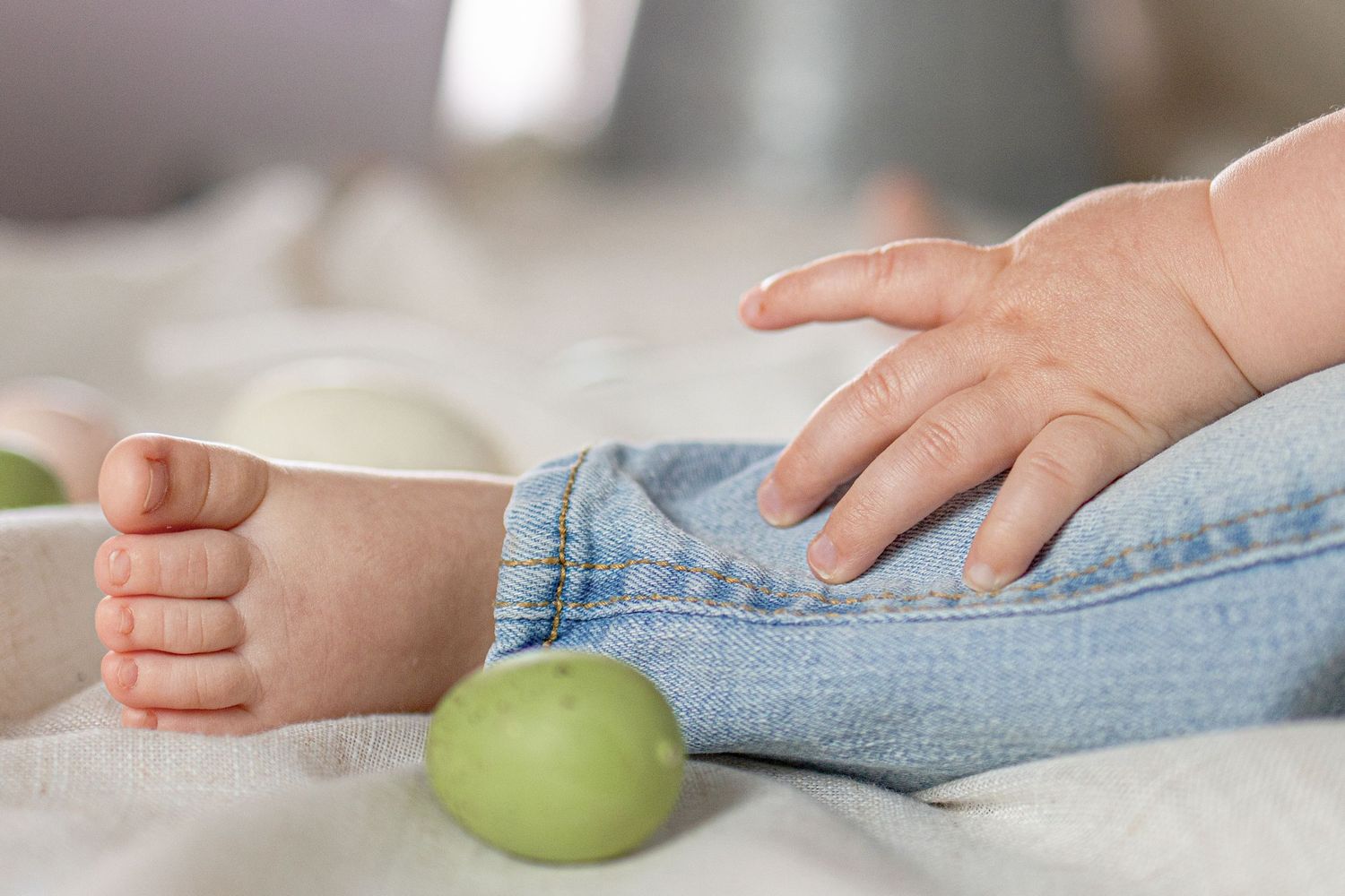 Close-up of baby hands and feet in denim jeans with a green apple on a soft white blanket.