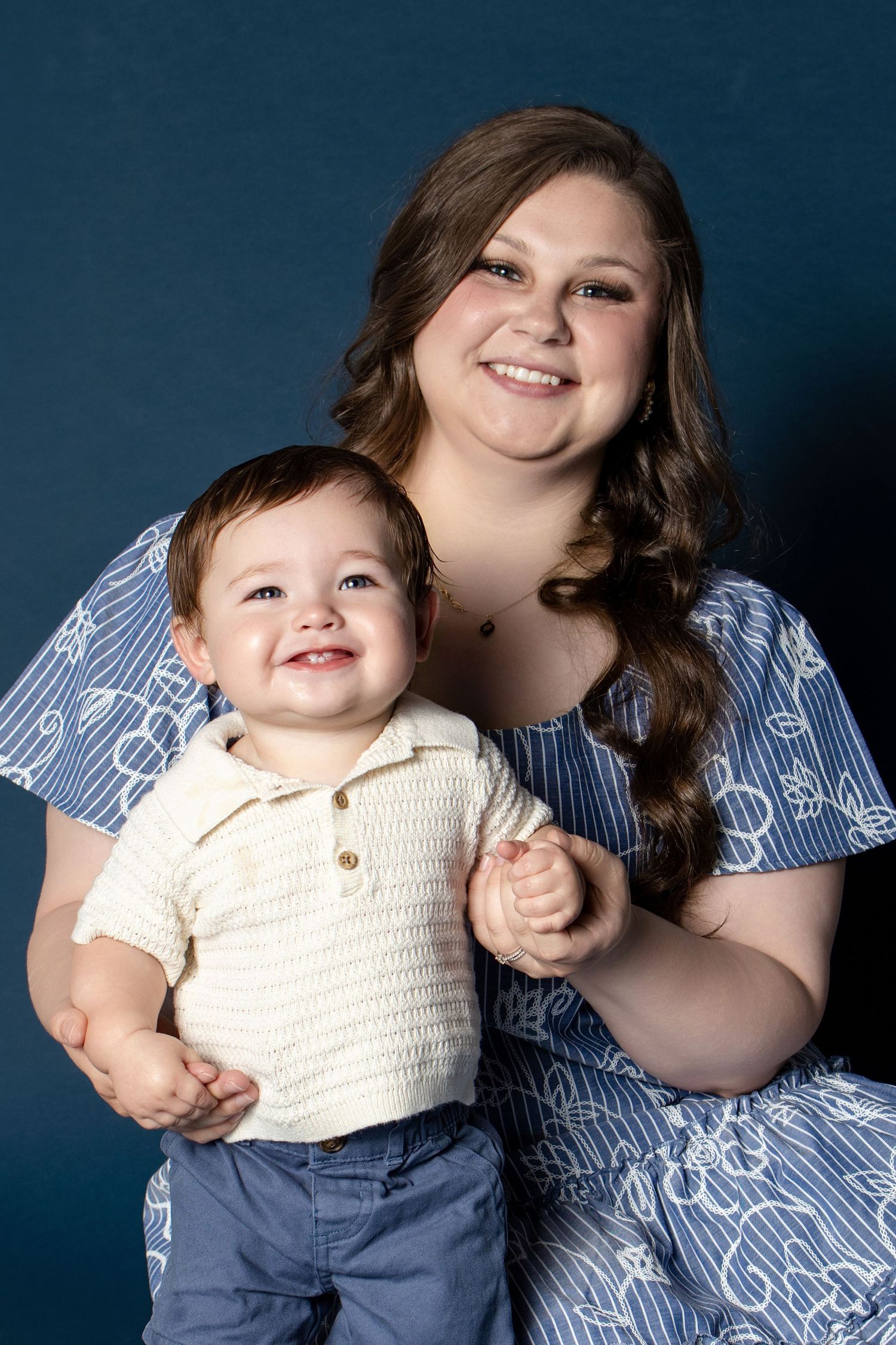 A smiling young woman in a blue floral dress poses with a happy toddler boy in a cream shirt for a studio portrait.