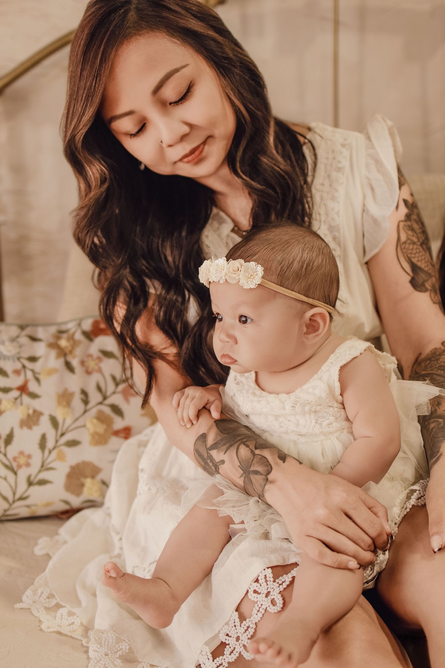 A mother with long dark hair and tattoos holds her baby girl in a white lace dress with floral headband.