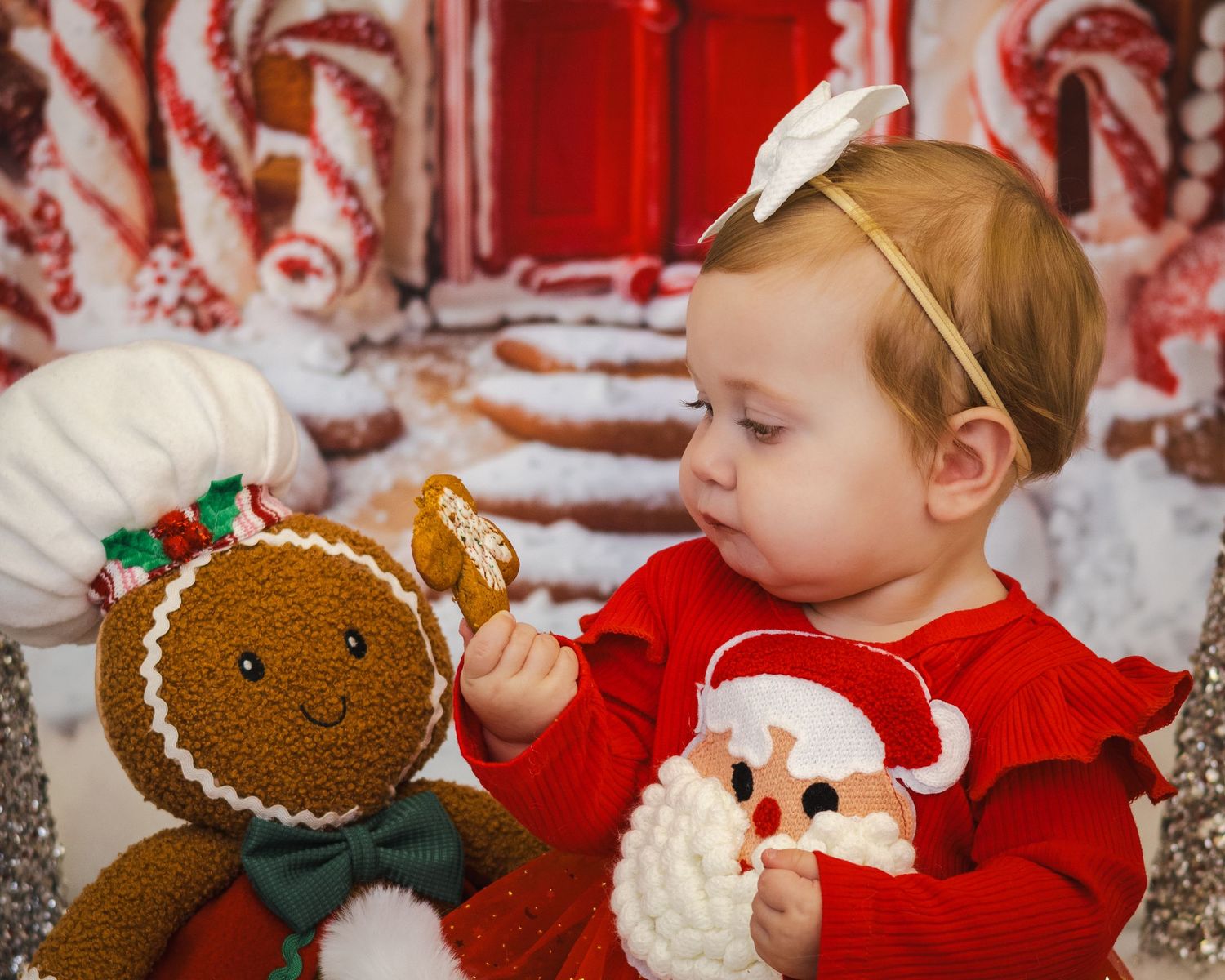 Baby girl in red dress holds gingerbread cookie next to plush gingerbread man in festive Christmas holiday photo.