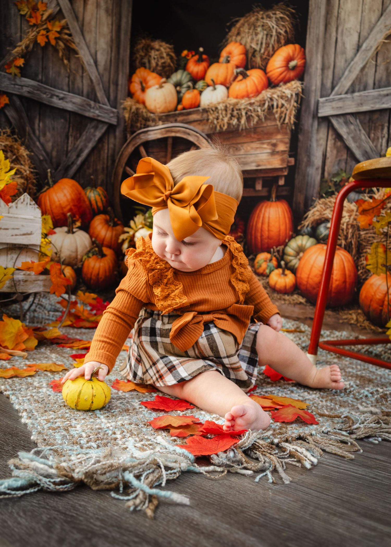 Baby girl in orange bow and plaid skirt sits among fall pumpkins and autumn leaves in rustic barn setting.