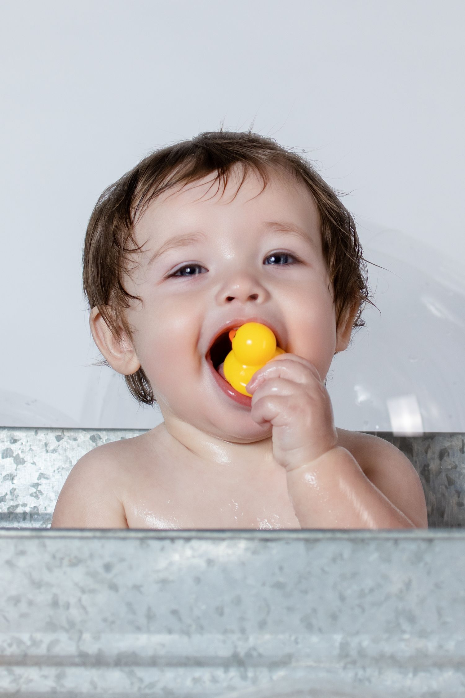 Baby in galvanized tub chewing on yellow rubber duck during bath time.