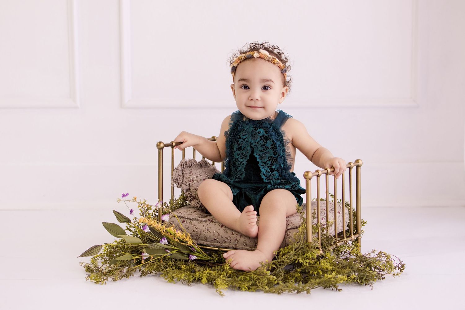 Baby girl in teal romper and floral headband sits on a gold mini bed surrounded by greenery in a studio photo.