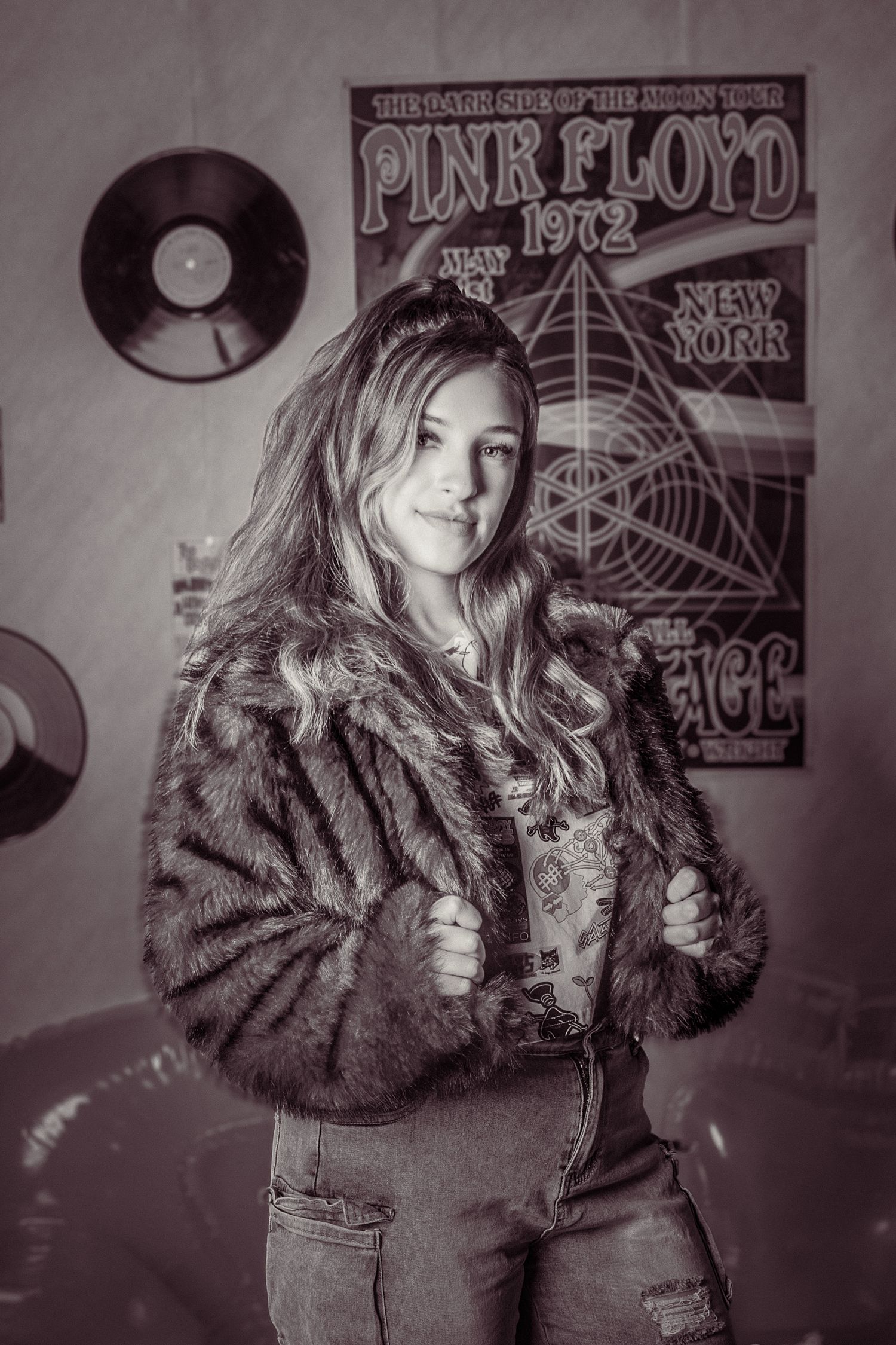 A young woman in a fur jacket poses confidently before a Pink Floyd 1972 concert poster in black and white.