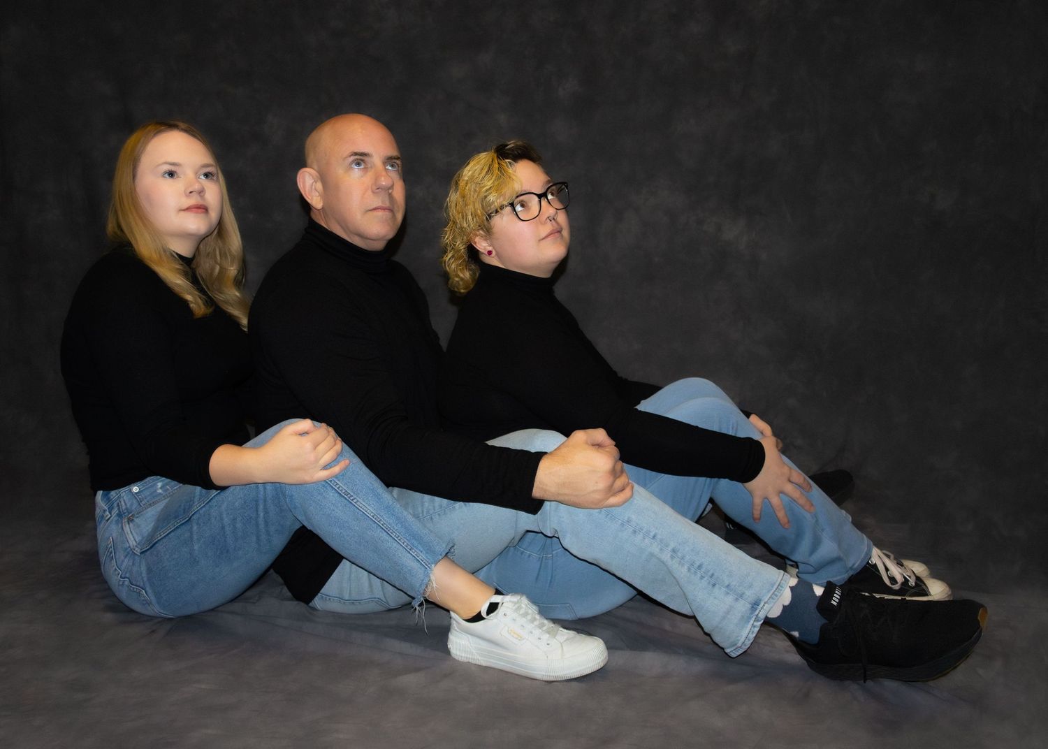 Three people in black tops and jeans sit together against a gray backdrop in a casual studio portrait photo.