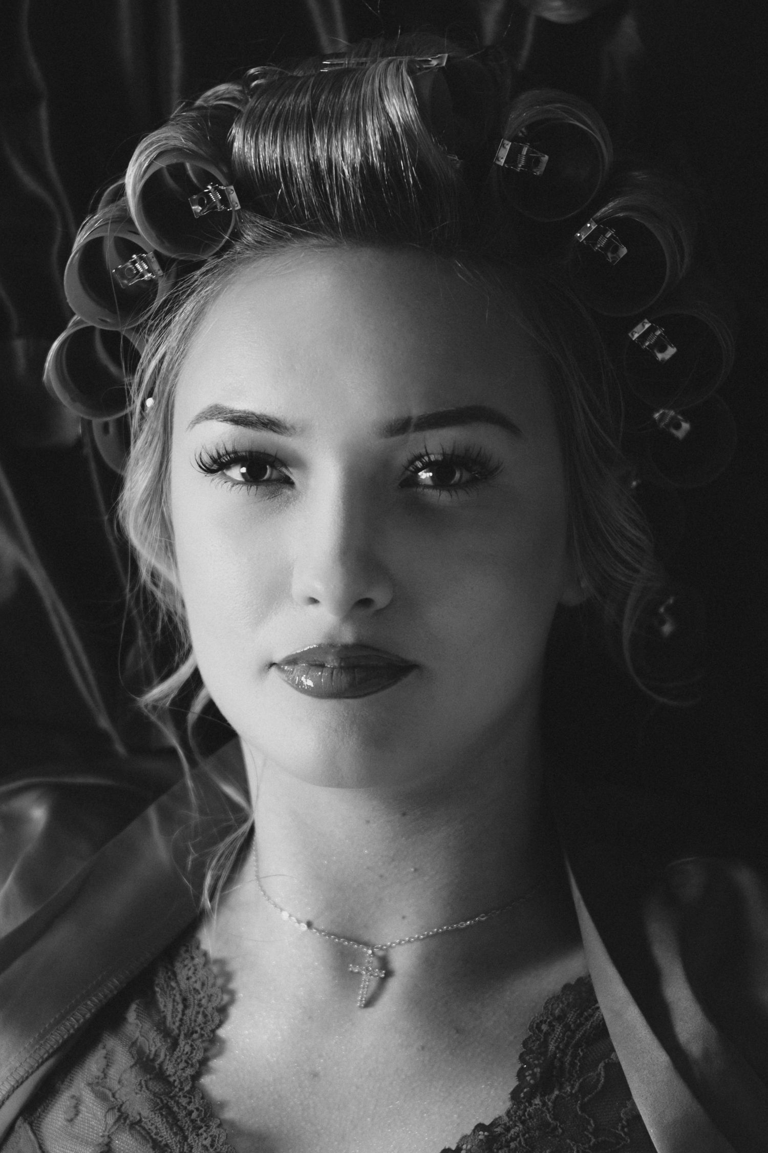Black and white close-up portrait of a woman with hair curlers and lace necklace looking at camera.