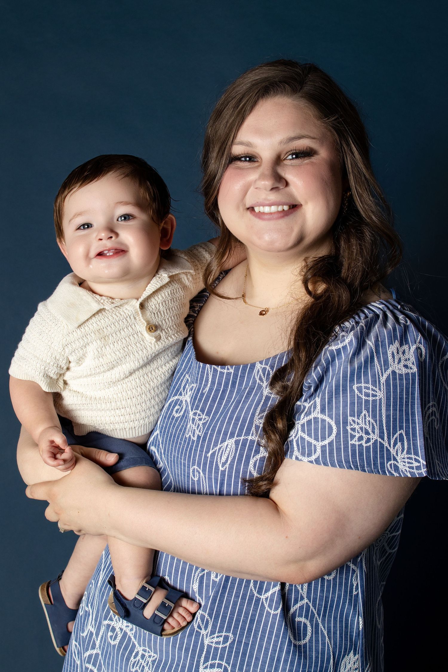 Smiling young woman in blue floral dress holding a happy baby boy in cream knit outfit against dark blue background.