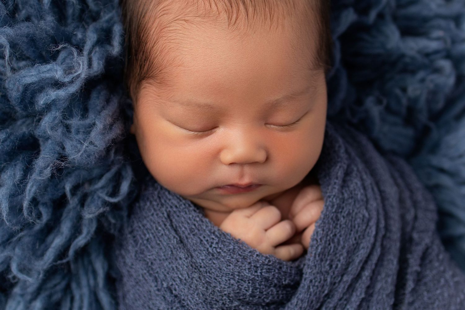 Peaceful newborn baby sleeping wrapped in a soft blue knit blanket on a fluffy blue textured backdrop.