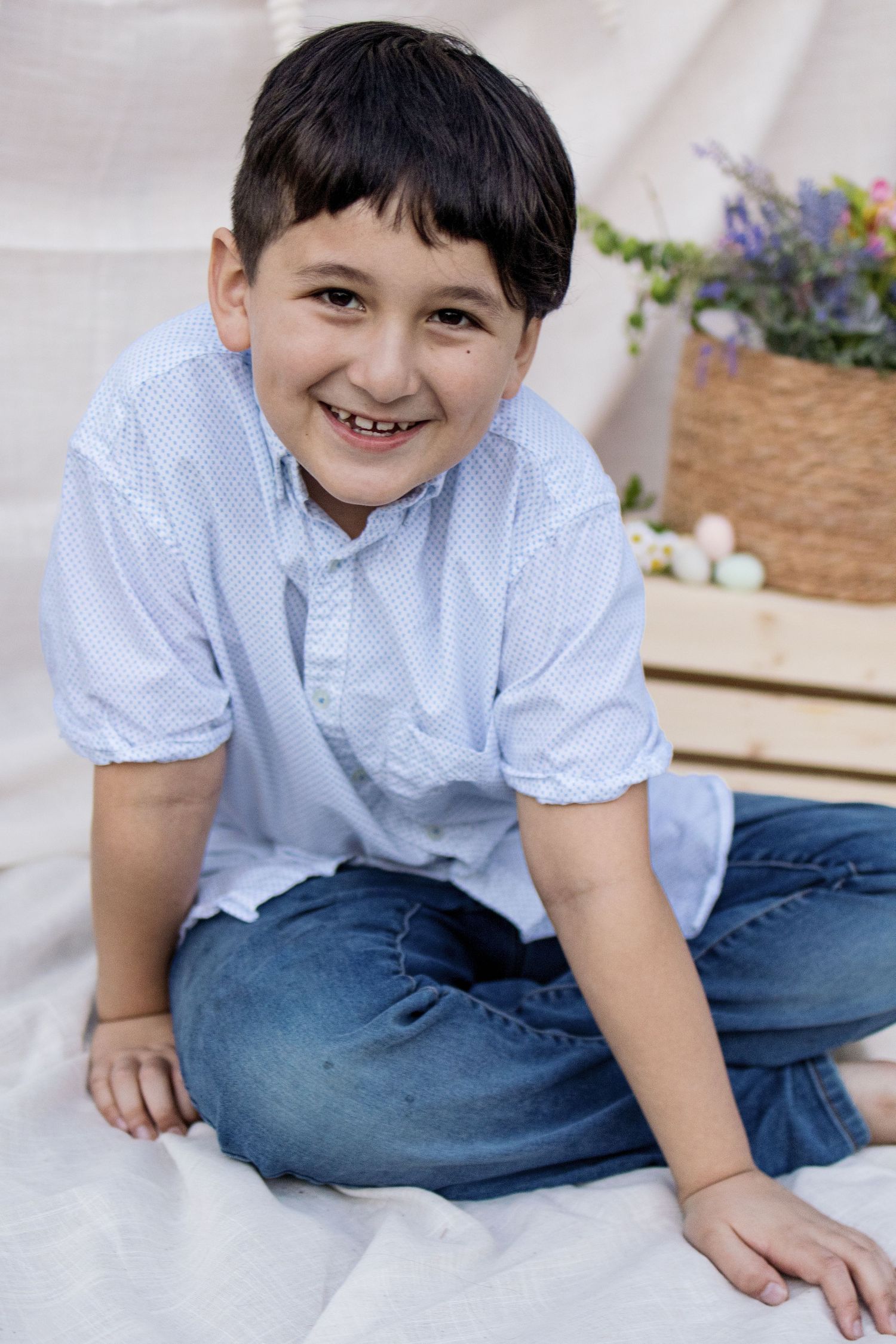A smiling boy in a light blue shirt and jeans sits cross-legged on a white blanket near Easter eggs.