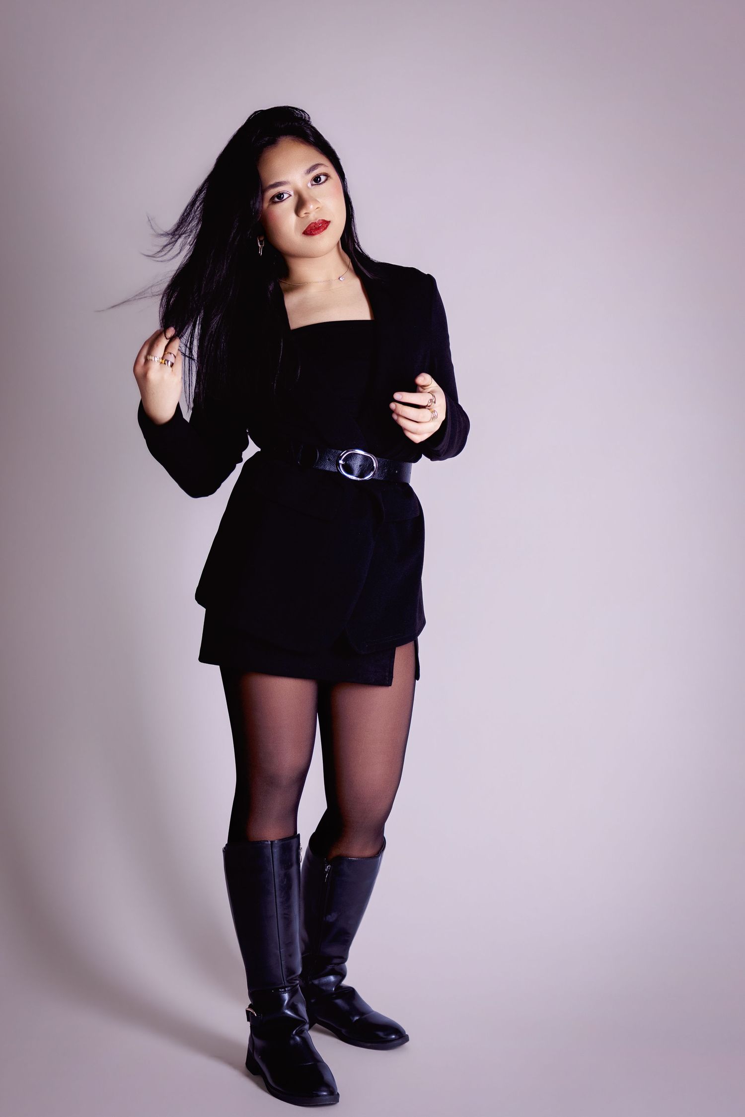 Young Asian woman in black mini dress, belt, tights, and knee-high boots posing in studio.