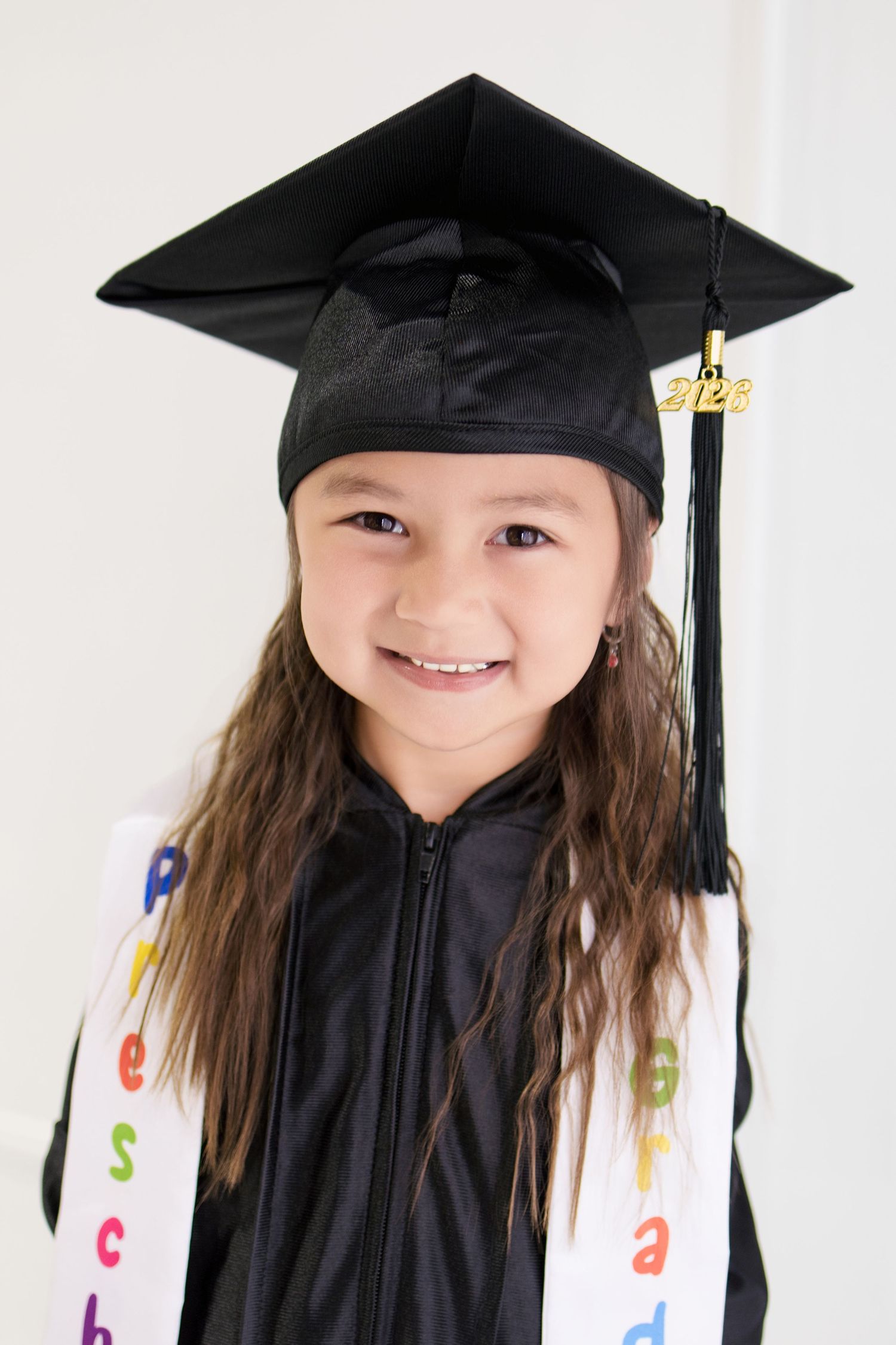 Cute young girl in black preschool graduation cap and gown with colorful stole smiling.